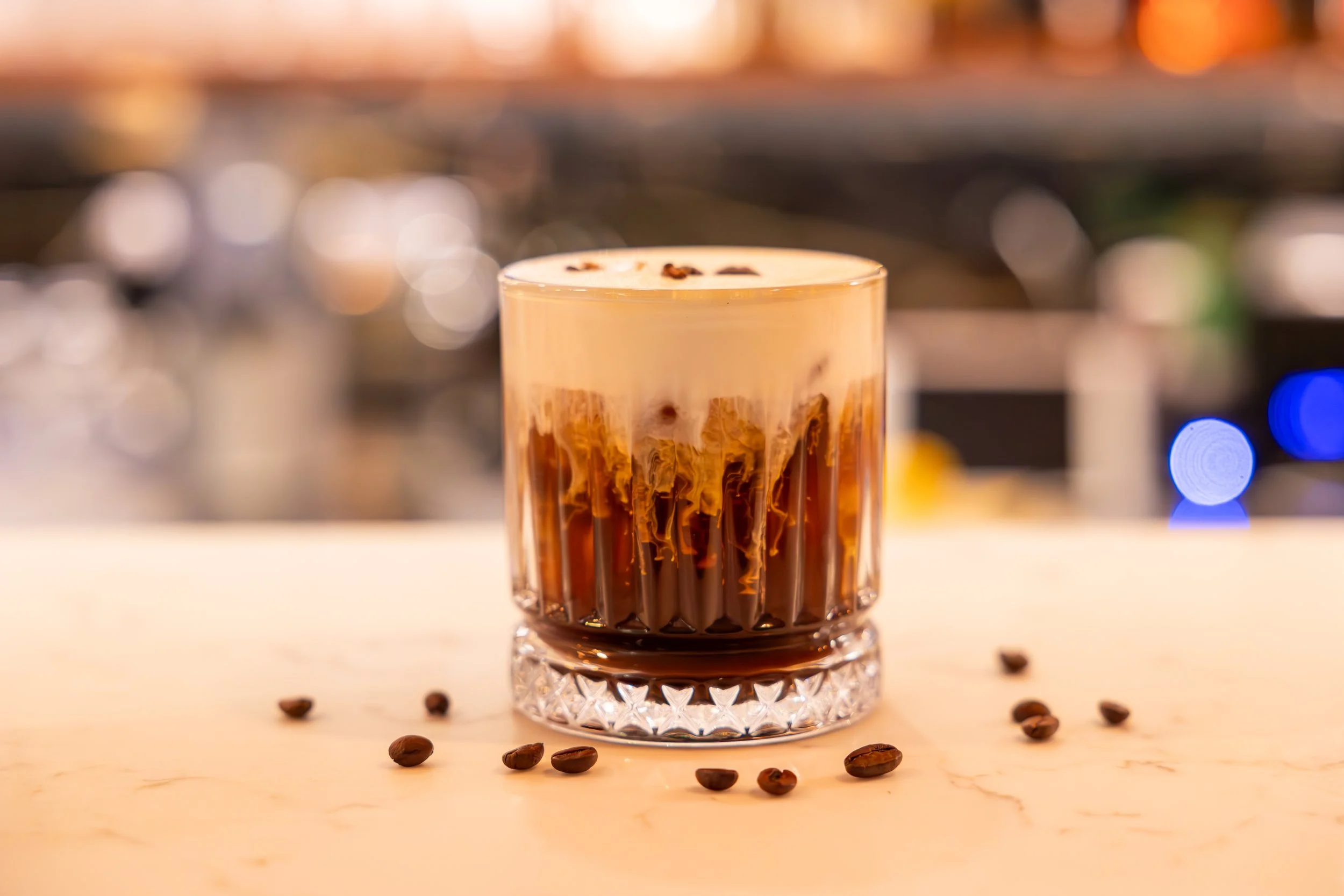 A tall glass with a layered iced coffee drink, topped with foam and coffee beans, on a light-colored surface with scattered coffee beans. Blurred background with warm lighting.