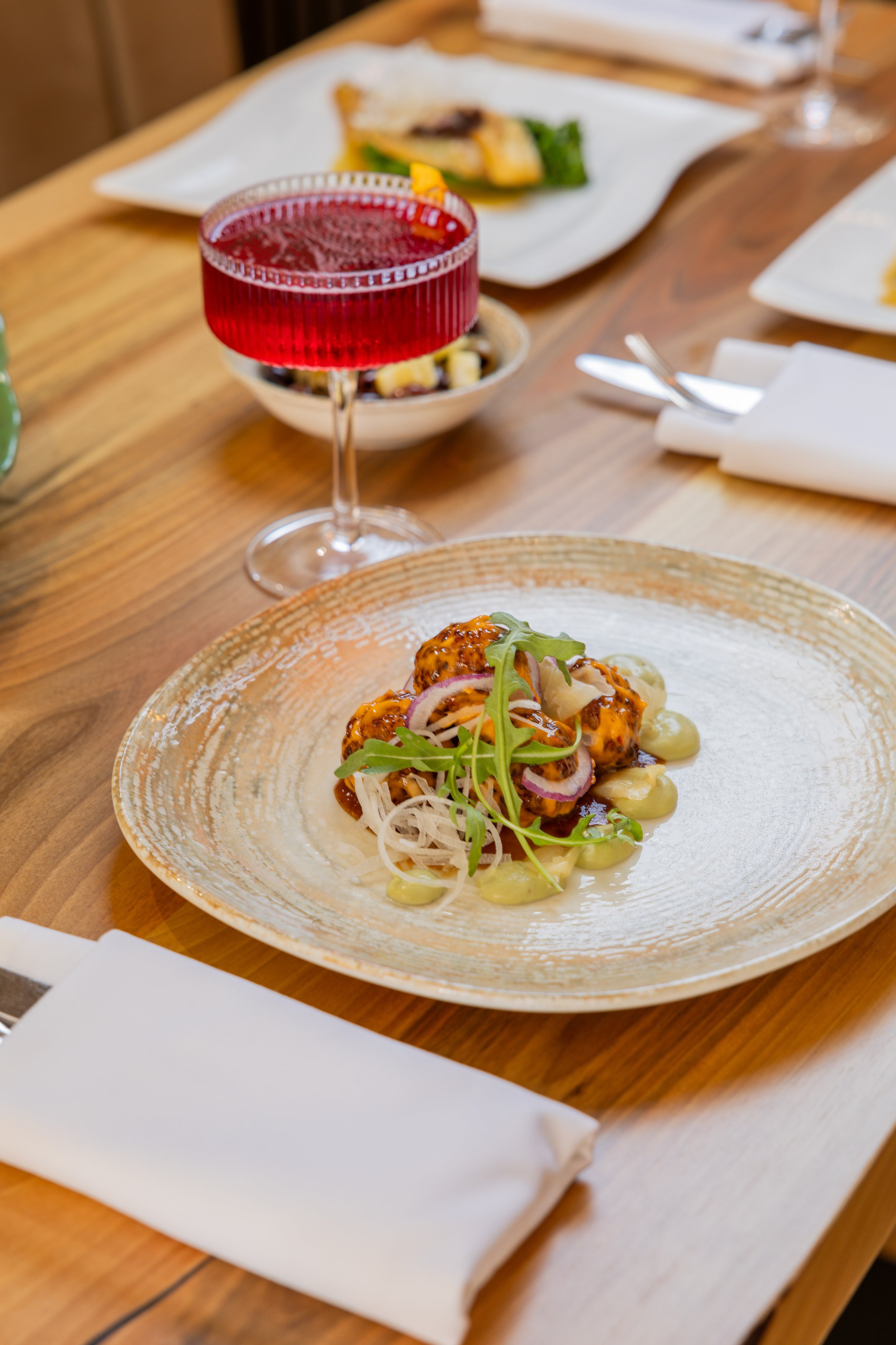 A dining table with a plate of food, a glass of red drink, and some plates in the background. The main dish appears to be a plated appetizer or entrée with greens, vegetables, or a salad.