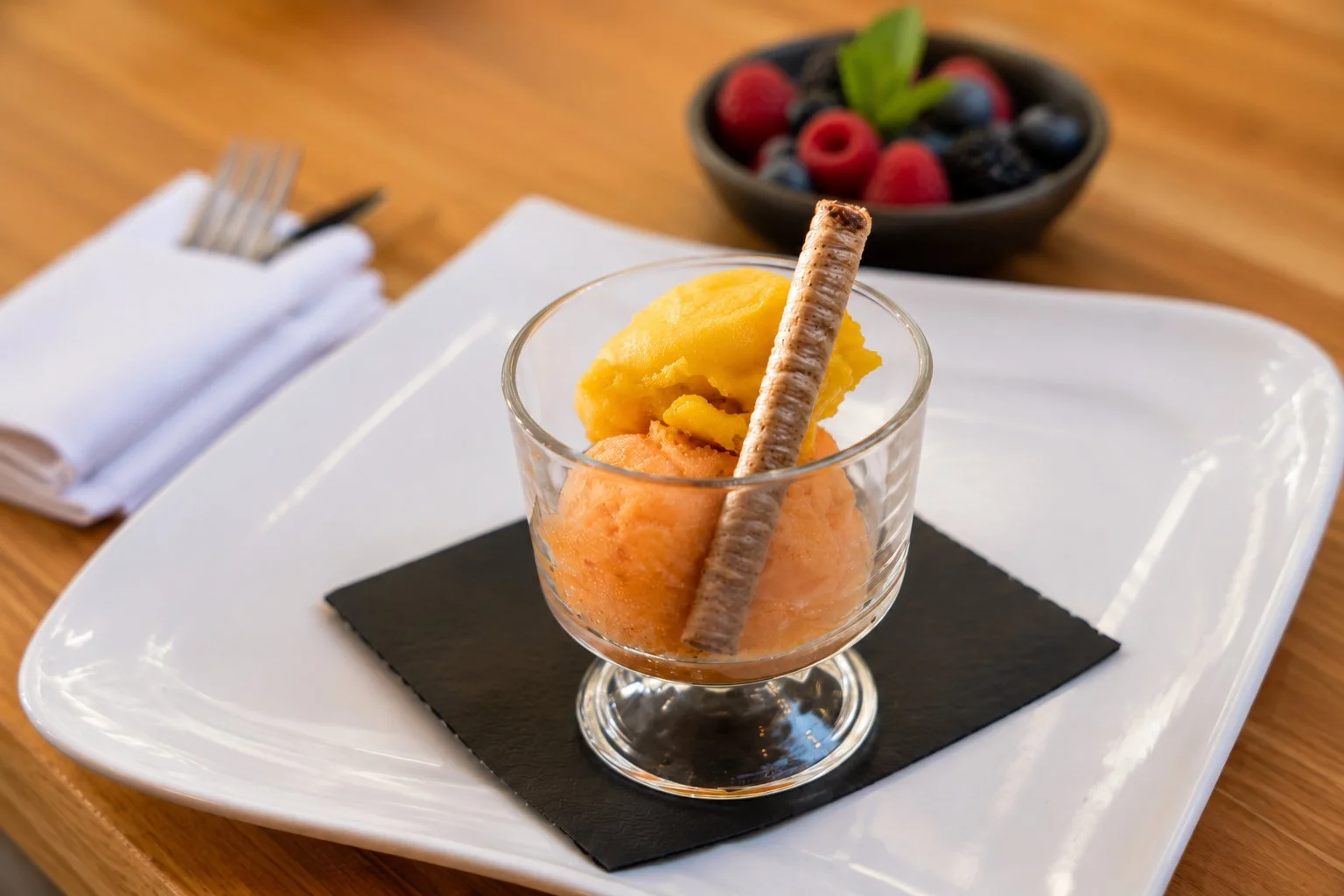A glass of two scoops of fruit-flavored sorbet, one yellow and one orange, garnished with a rolled wafer cookie, placed on a black napkin on a white plate. In the background, a bowl of assorted berries, including raspberries, blackberries, and blueberries, with a green mint leaf, on a wooden table.