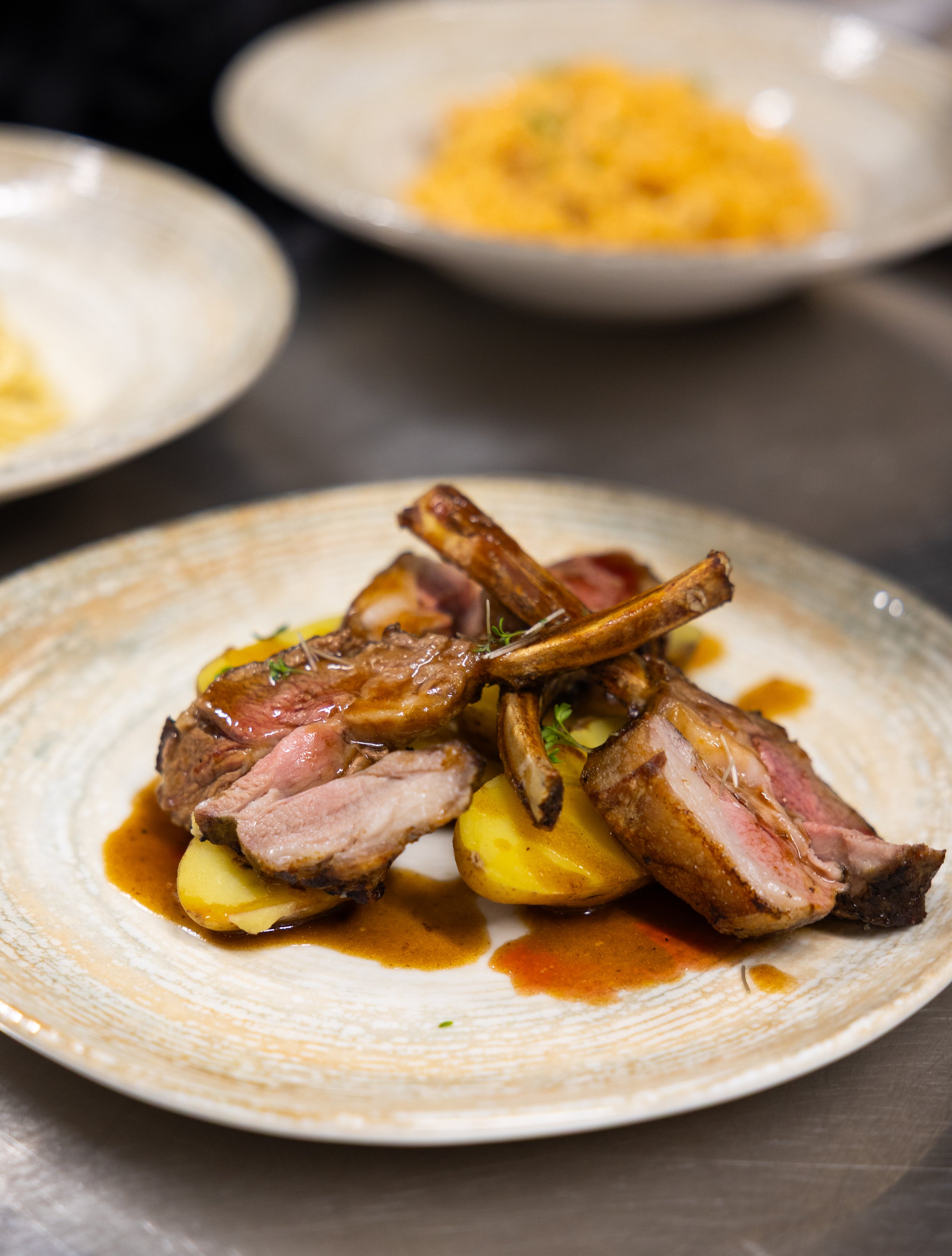 A plate of cooked lamb chops with sauce and potatoes, with two blurred dishes of side dishes in the background.