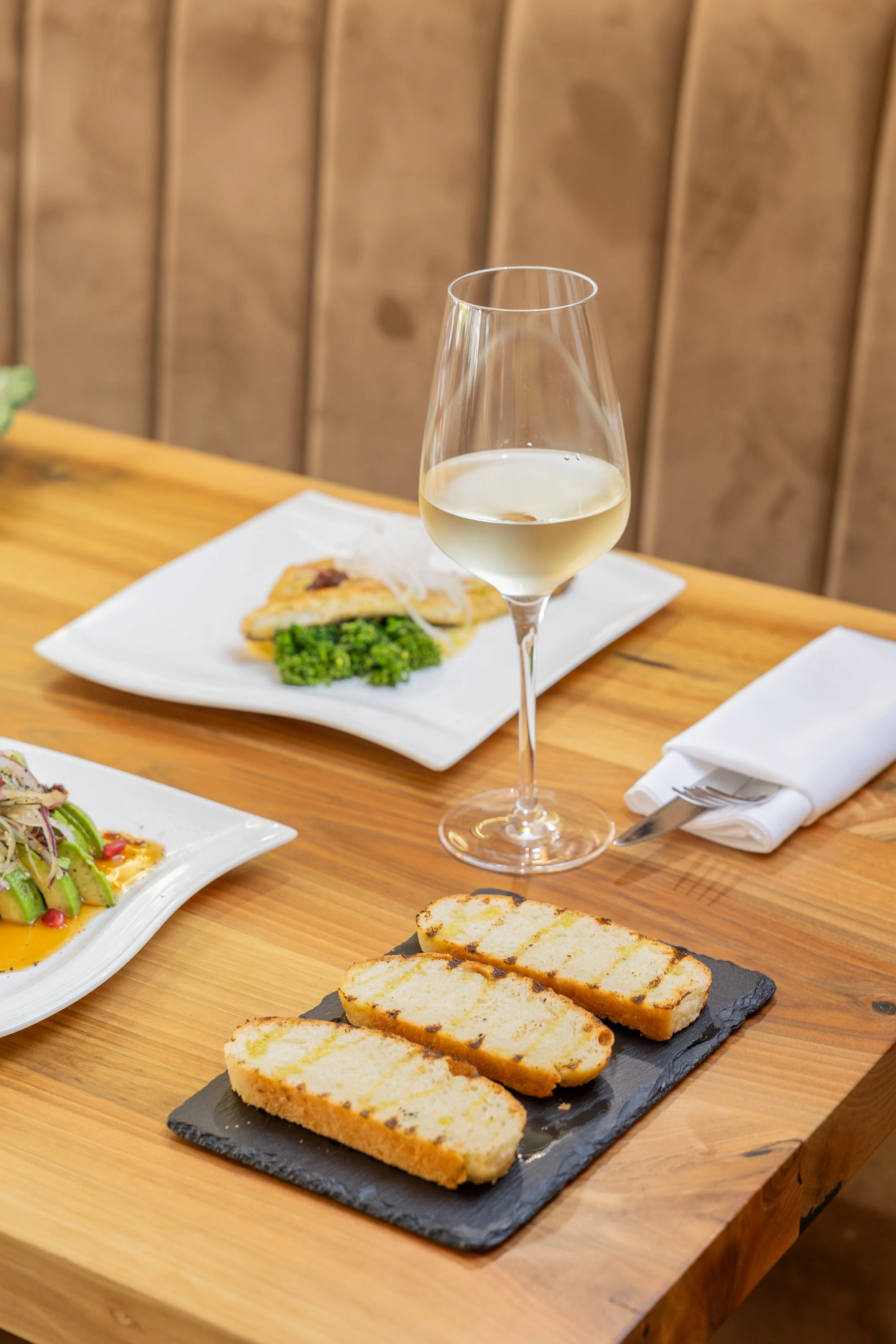 A glass of white wine on a wooden table with various plates of food, including grilled bread, vegetables, and fish, in a restaurant setting with wooden paneling in the background.