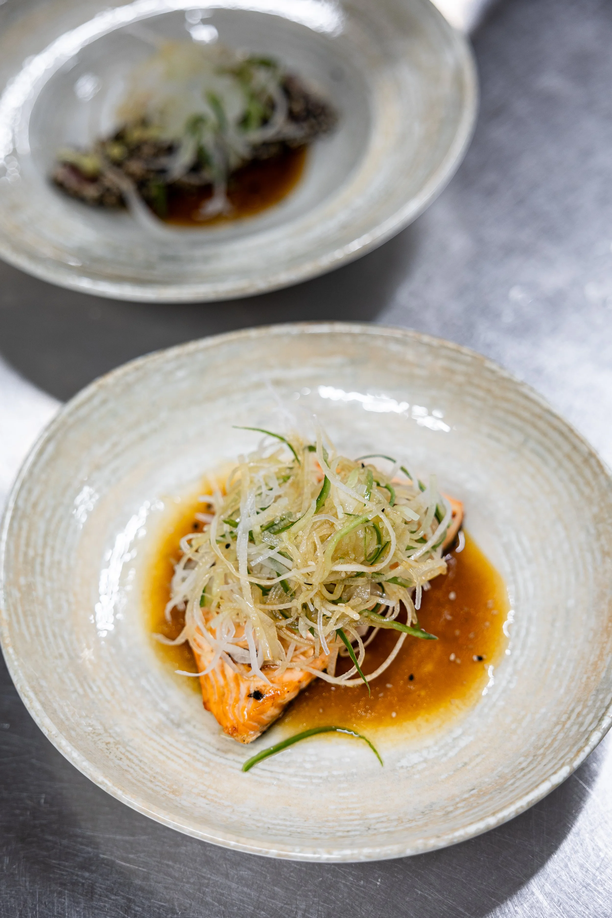 A dish featuring grilled salmon topped with shredded vegetables and served with a sauce in a beige ceramic bowl.