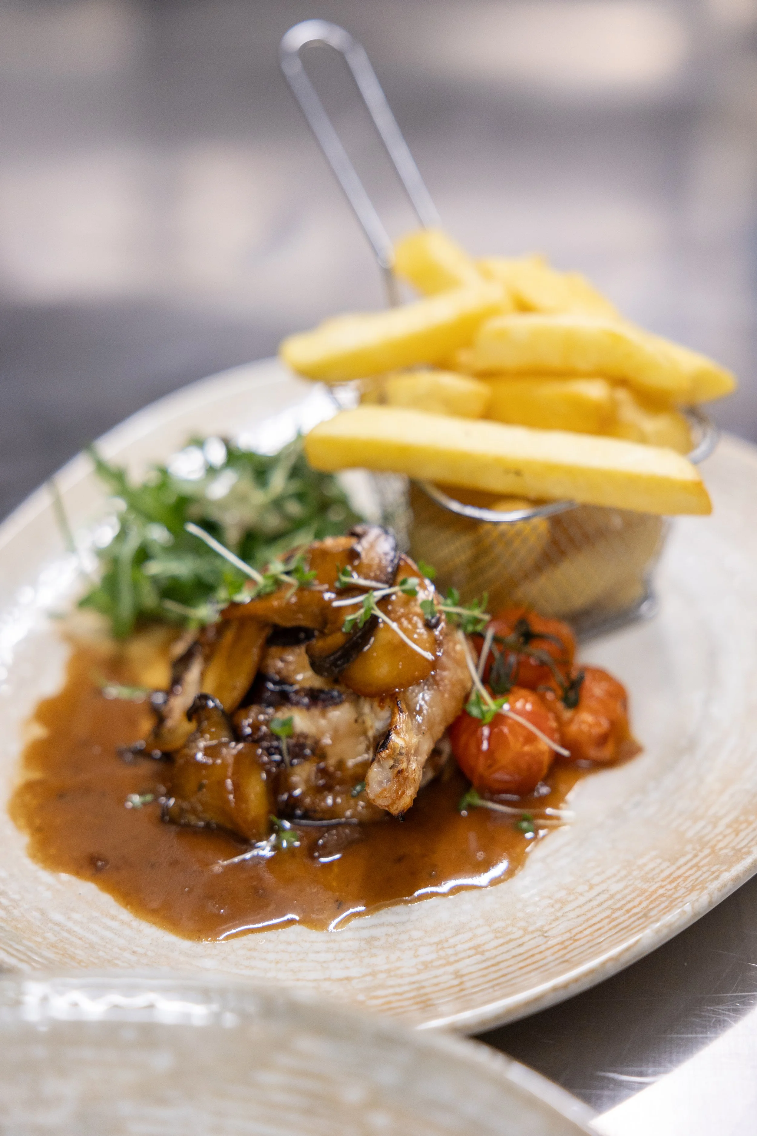 Plate with grilled chicken topped with mushroom gravy, French fries in a metal basket, green garnish, and roasted vegetables.