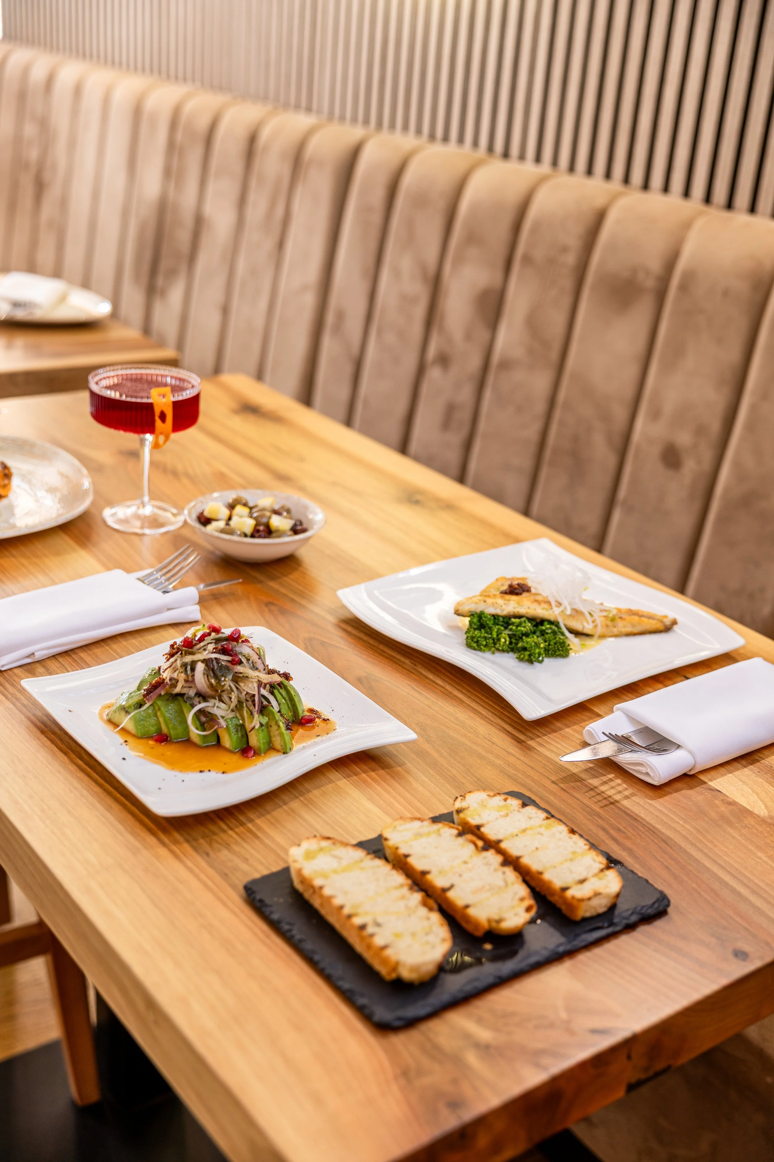 A wooden dining table set with plates of Asian-inspired dishes, a glass of red beverage, and a bowl of salad, with beige cushioned seating in the background.