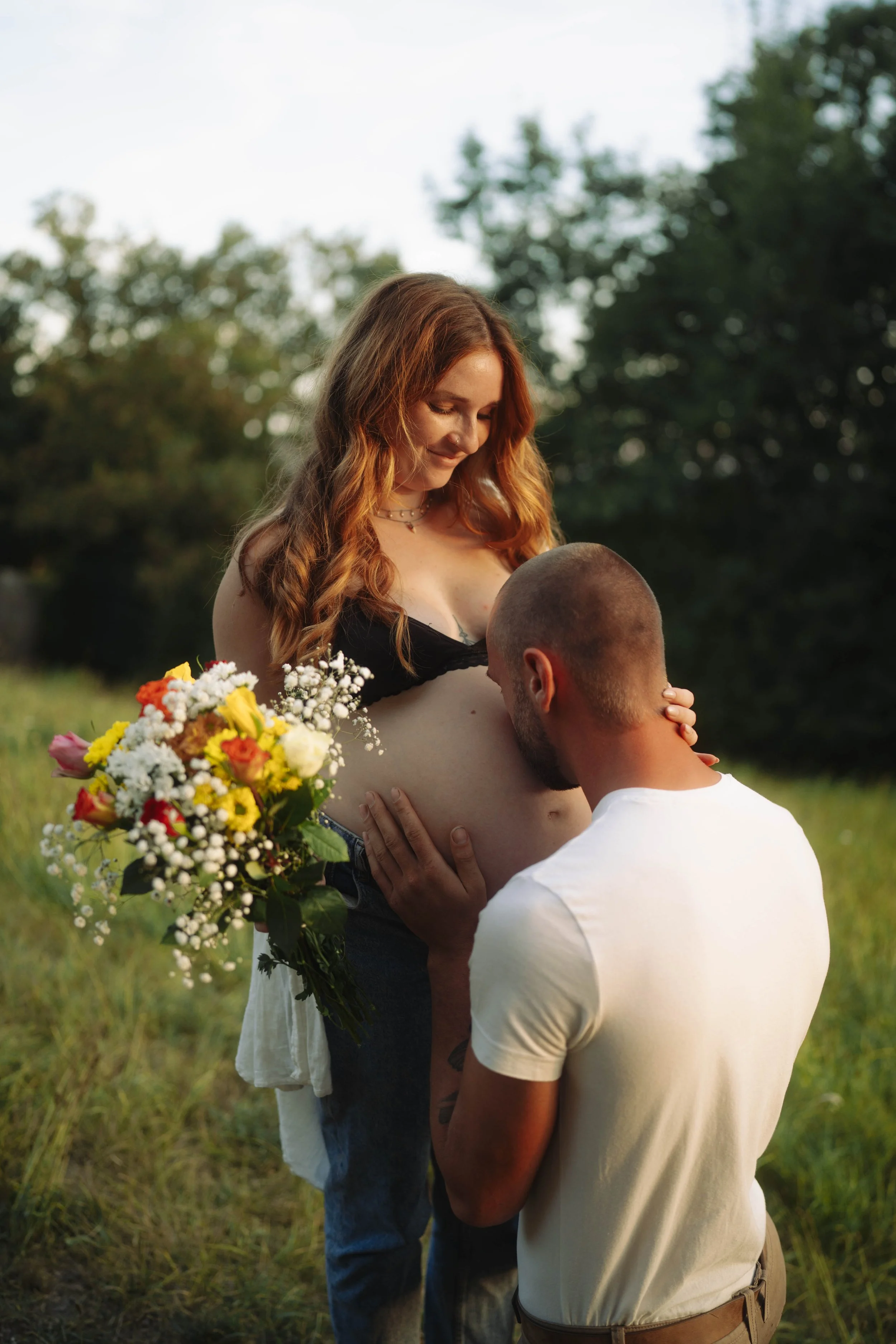Ein Paar in einer Wiese bei Sonnenuntergang, der Mann ist auf Kniehöhe und hält das Baby, die Frau steht mit einem Blumenstrauß.
