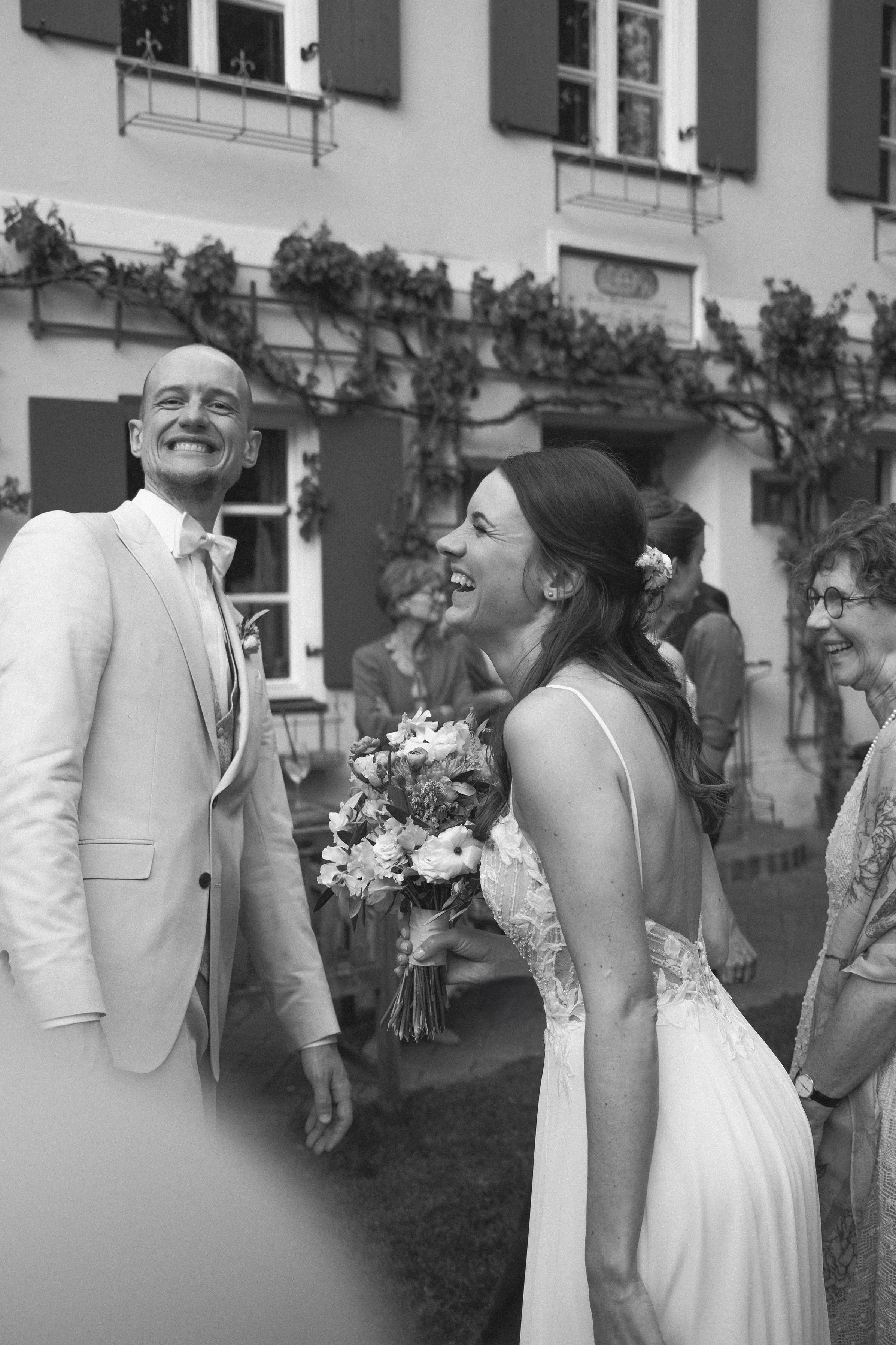Ein glückliches Brautpaar bei einer Hochzeit, das sich gegenseitig anschaut, während sie Lachen. Der Bräutigam trägt einen Anzug, die Braut ein Brautkleid und hält einen Blumenstrauß. Im Hintergrund sind weitere Gäste und eine weiße Hausfassade mit Fensterläden.