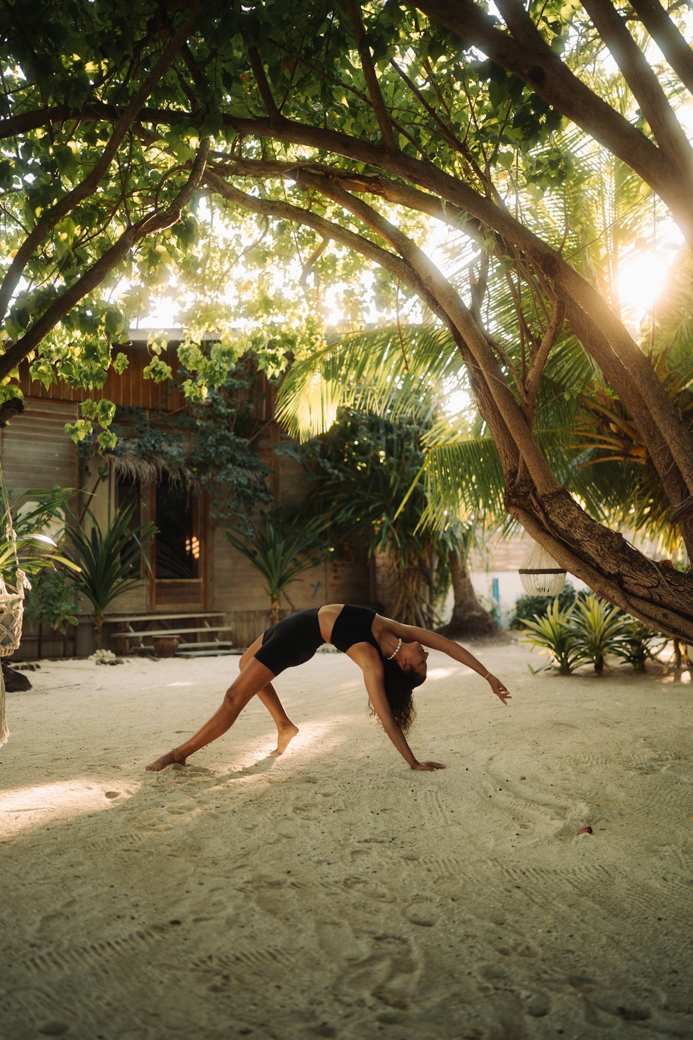 Eine Frau macht Yoga auf einem Sandboden im Garten unter einem Baum, Sonnenlicht scheint durch die Blätter.