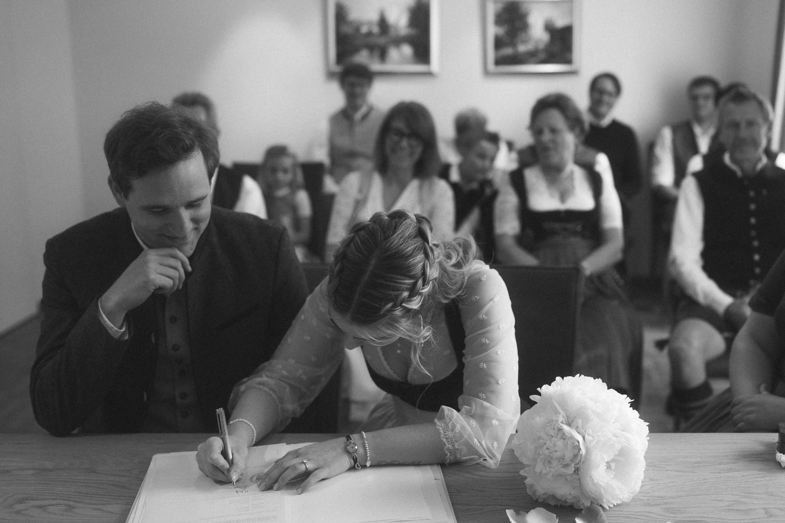 Ein Brautpaar unterschreibt einen Heiratsvertrag oder ein Dokument bei einer Hochzeitszeremonie, umgeben von Gästen in einem festlichen Raum mit Gemälden an der Wand.