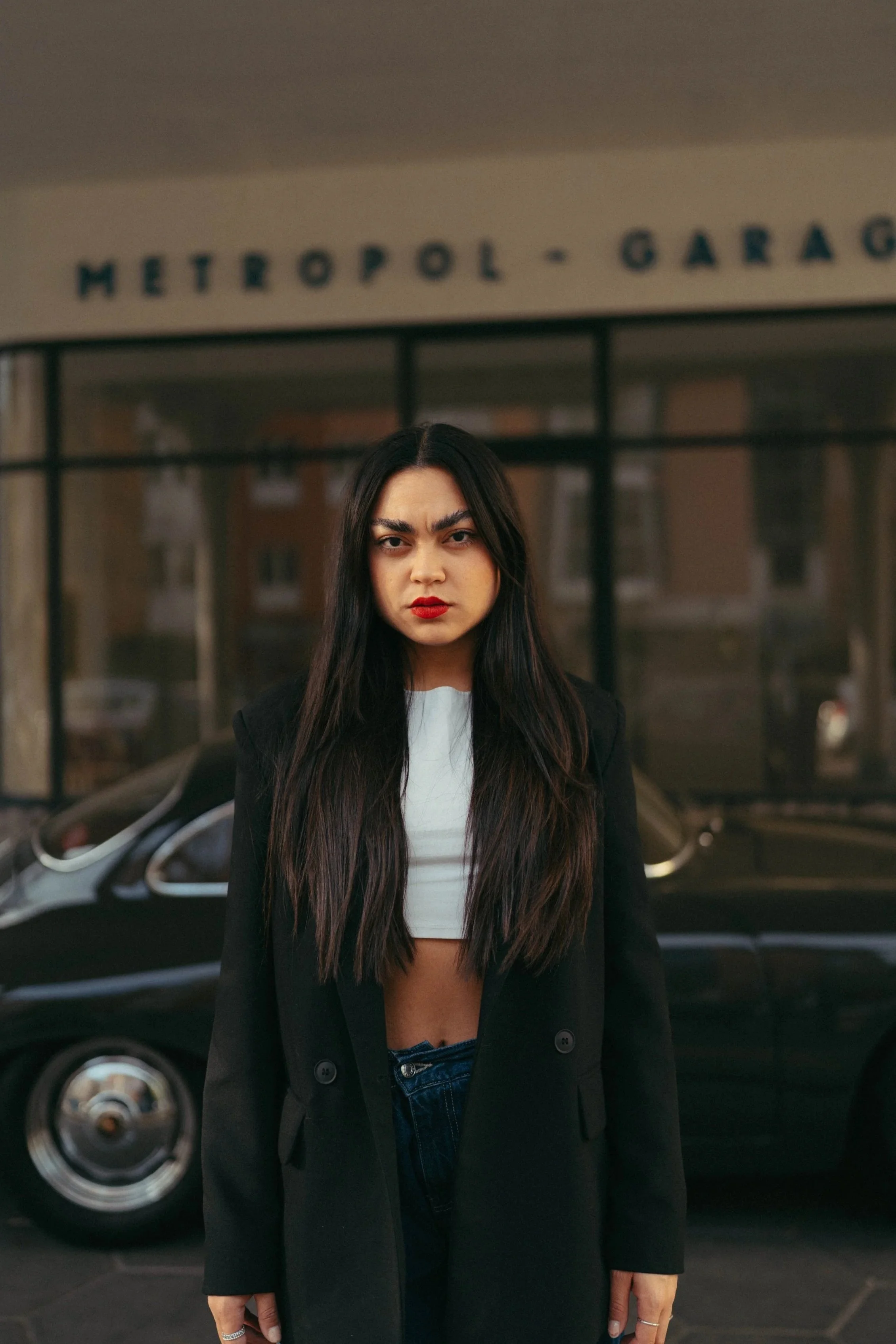 Junge Frau mit dunklem Haar, rotem Lippenstift und schwarze Jacke vor einem schwarzen Auto, im Hintergrund ein Gebäude mit Glasfenstern und einem Schild mit der Aufschrift 'Metropol-Garage'.