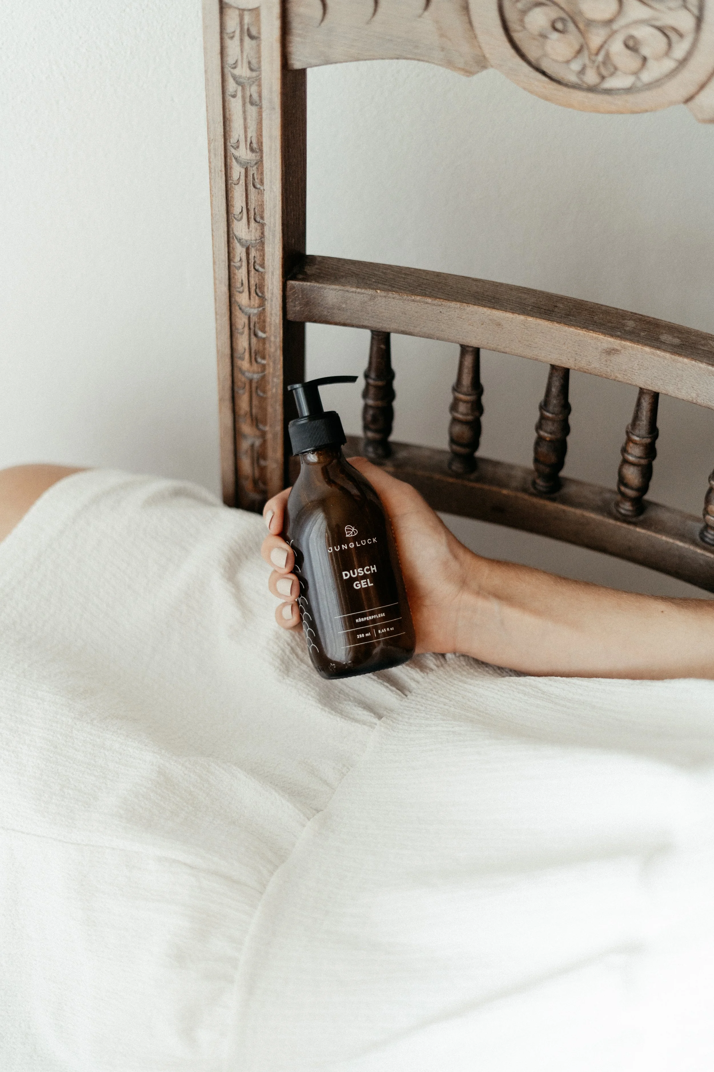 Person hält eine braune Flasche mit Duschgel in der Hand, während sie im Bett liegt, mit einem Holzkopfende im Hintergrund.