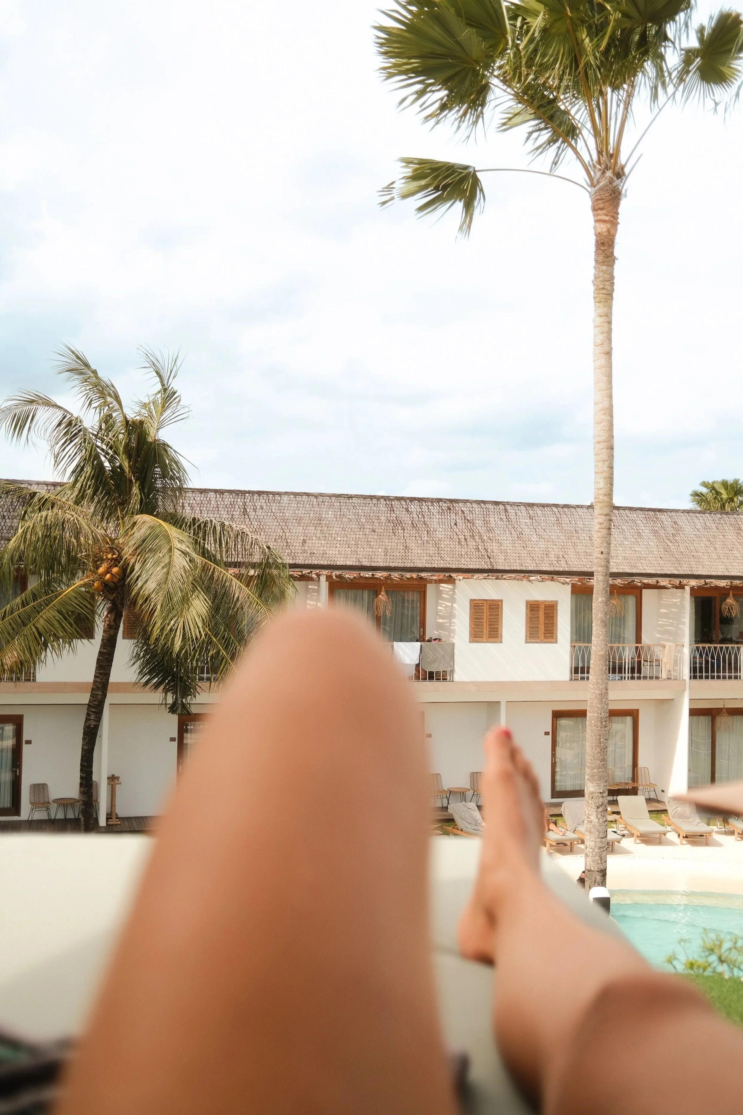 Blick auf einen Hotelkomplex mit weißen Wänden, mehreren Balkonen, Liegestühlen und Palmen im Vordergrund, Pool sichtbar, unter einem bewölkten Himmel.