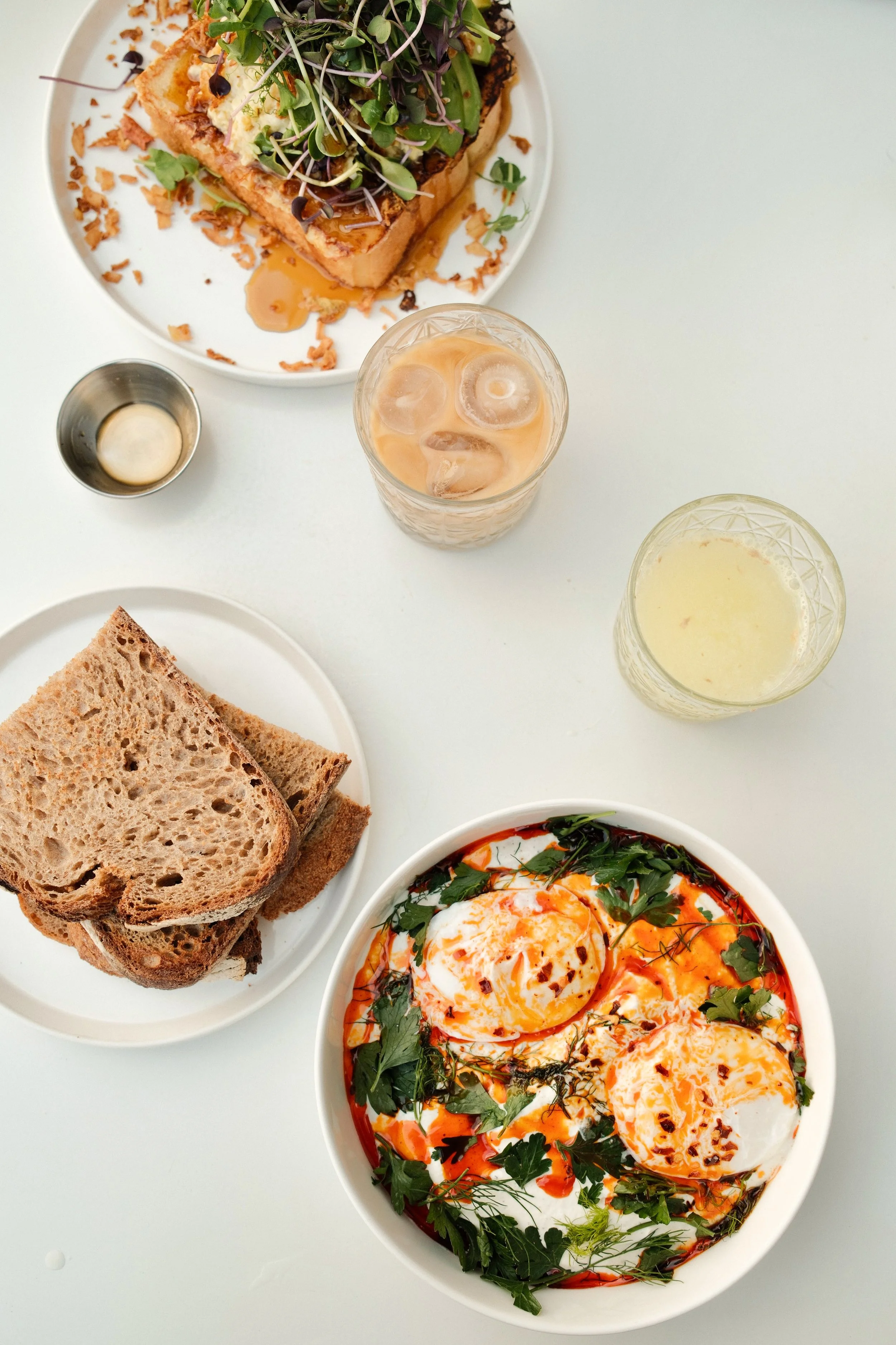 Bunte Mahlzeit mit ofenwarmen Eier, Tomaten, frischen Kräutern, Vollkornbrot, Salat, Joghurt, und Getränken auf weißem Tisch.