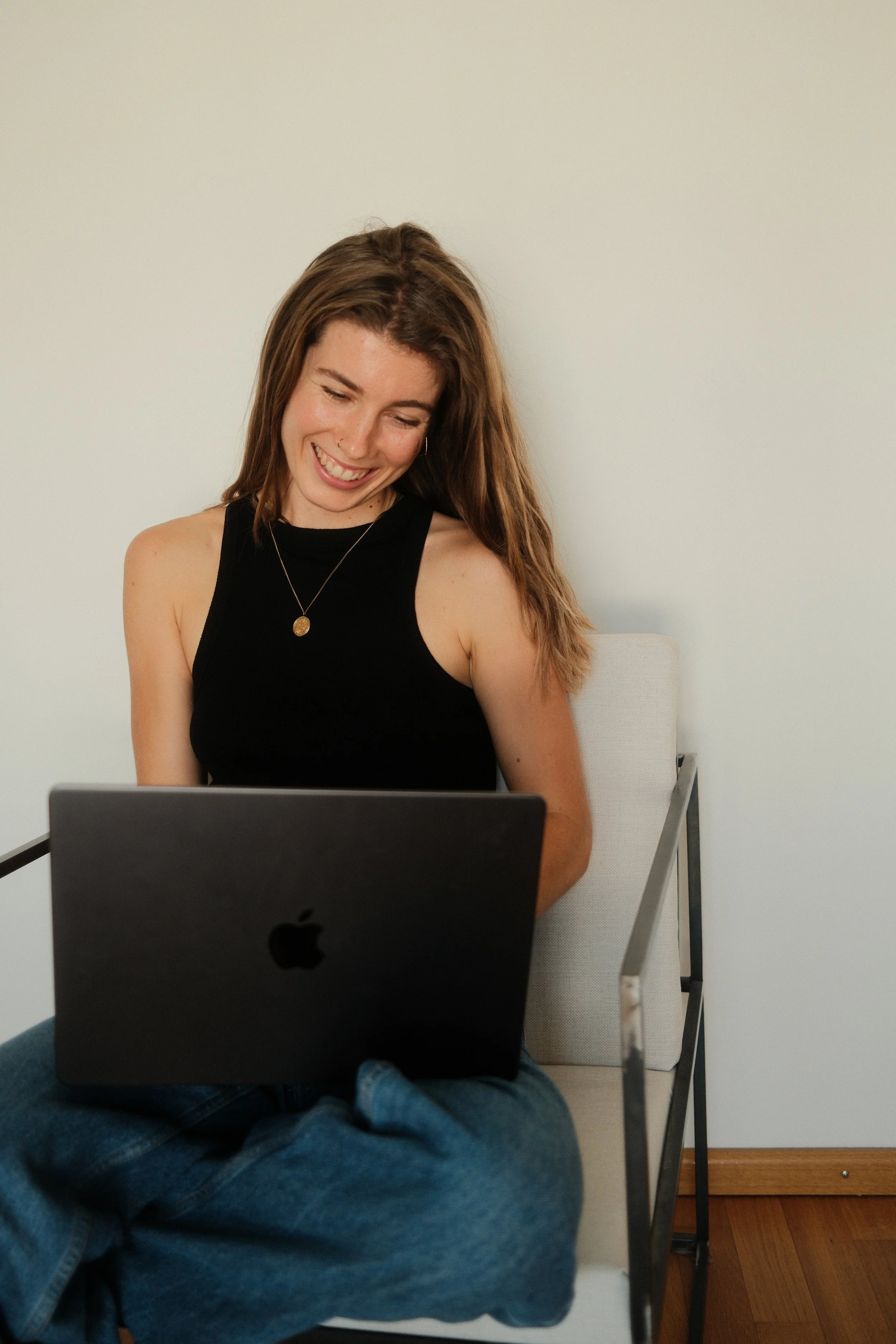 Junge Frau mit braunen Haaren, schwarzen Top und Goldkette sitzt an einem Tisch mit einem Laptop, lächelt und schaut nach unten. Sie ist in einem hellen Raum, Hintergrund eine weiße Wand.