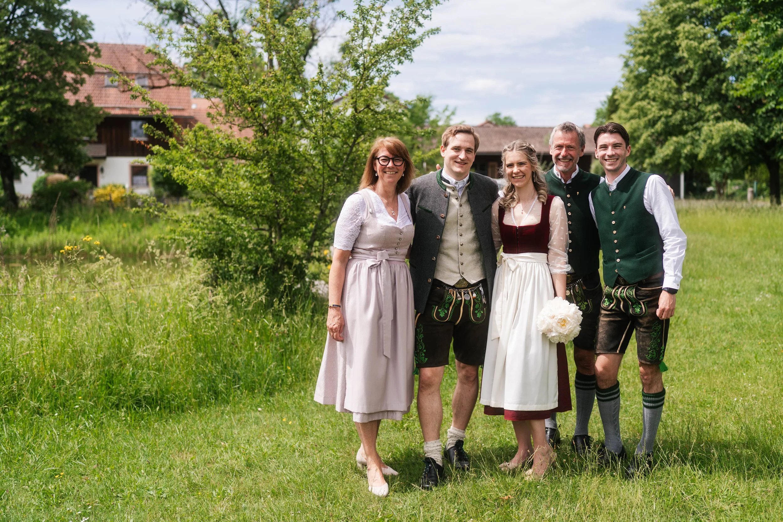 Eine Gruppe von fünf Menschen, darunter zwei Männer und drei Frauen, trägt traditionelle bayerische Kleidung und steht im Grünen, vermutlich bei einer Hochzeit außerhalb eines Hauses mit Garten, sonniges Wetter mit blauem Himmel.
