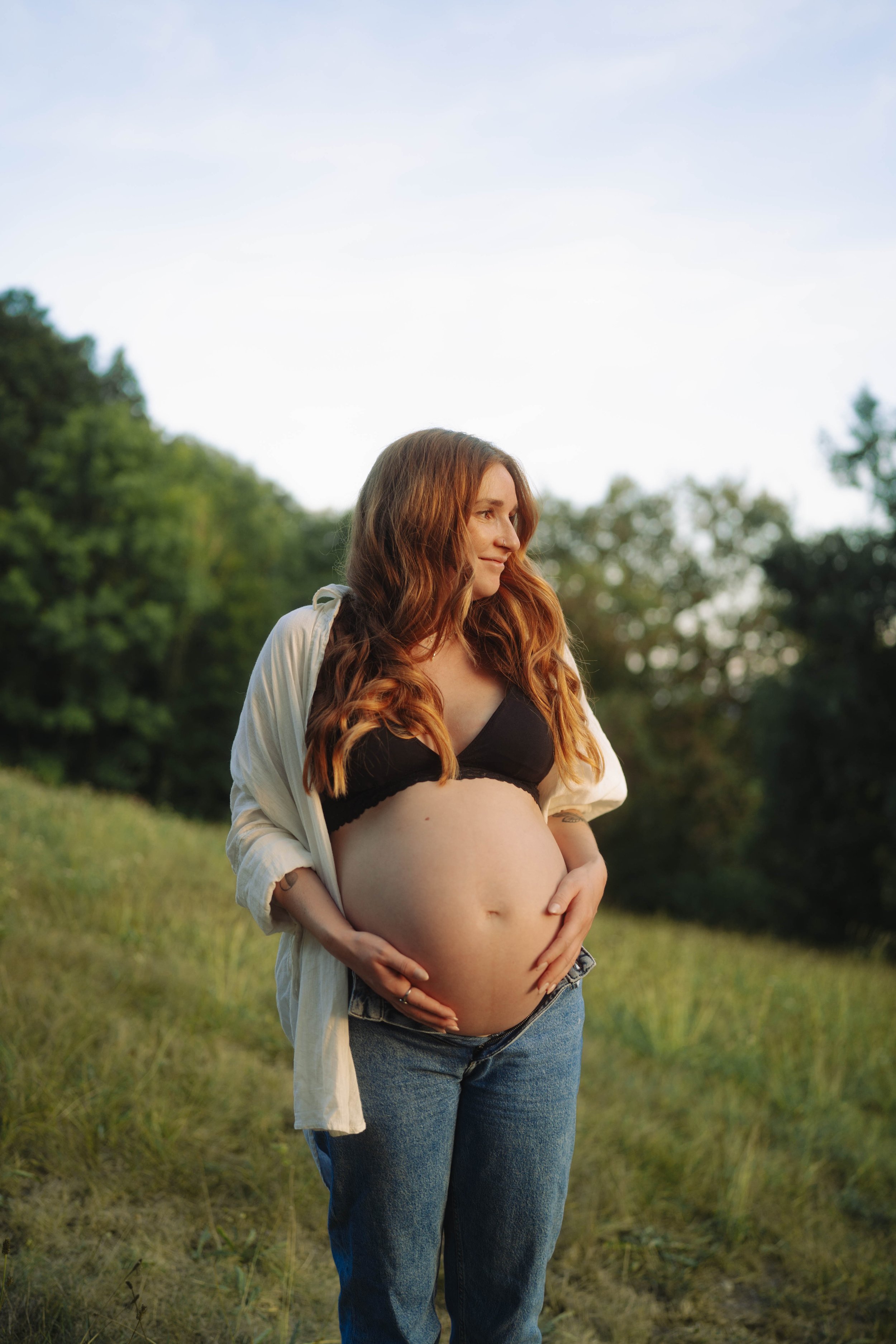 Schwangere Frau mit langen braunen Haaren positioniert ihre Hände auf ihren Bauch im Freien, umgeben von Bäumen und Gras bei Tageslicht.