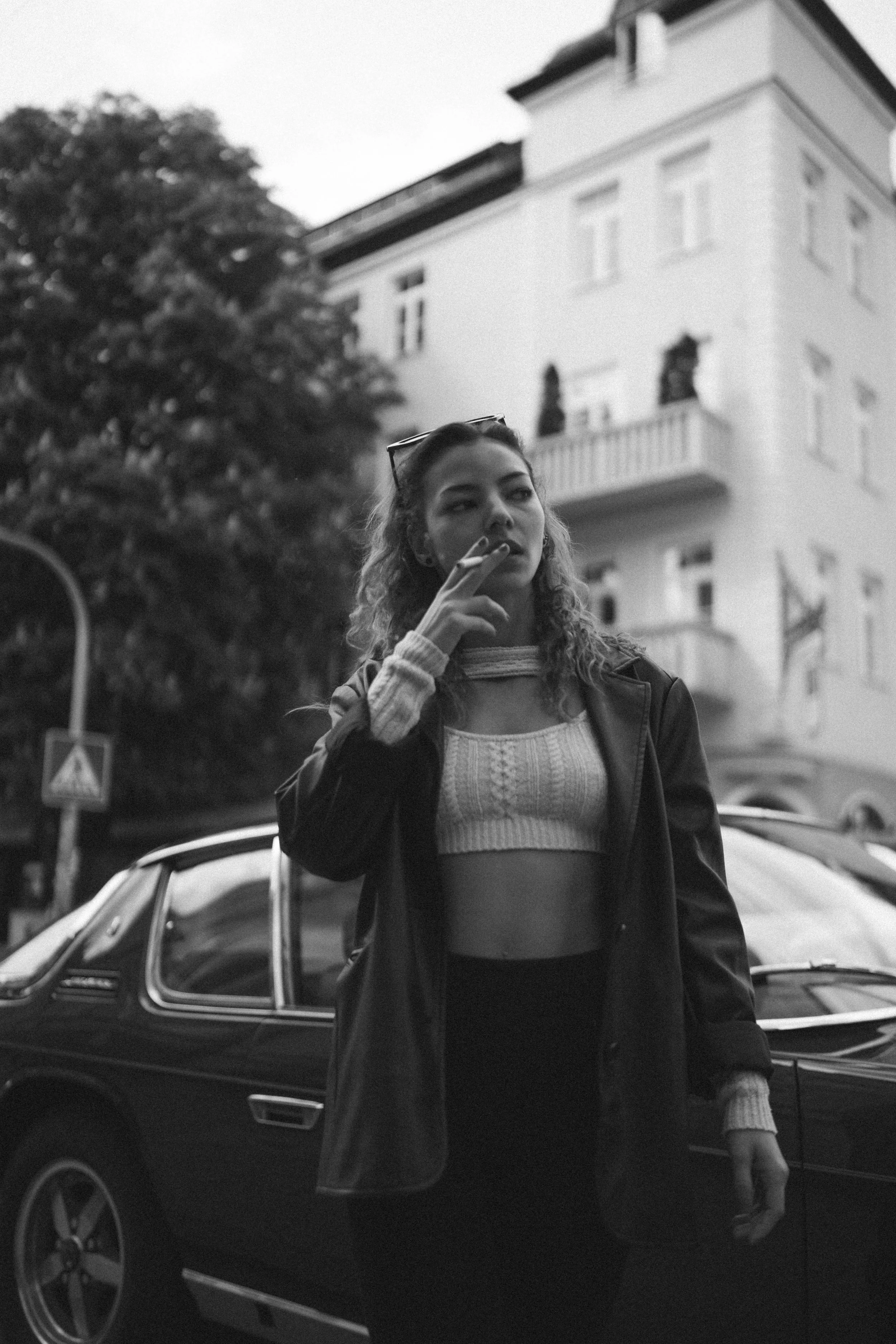 Schwarzes und weißes Foto einer jungen Frau, die draußen vor einem Auto steht, mit einer Zigarette in der Hand, Sonnenbrille auf dem Kopf, an einem Tag mit mehrstöckigem Gebäude im Hintergrund.