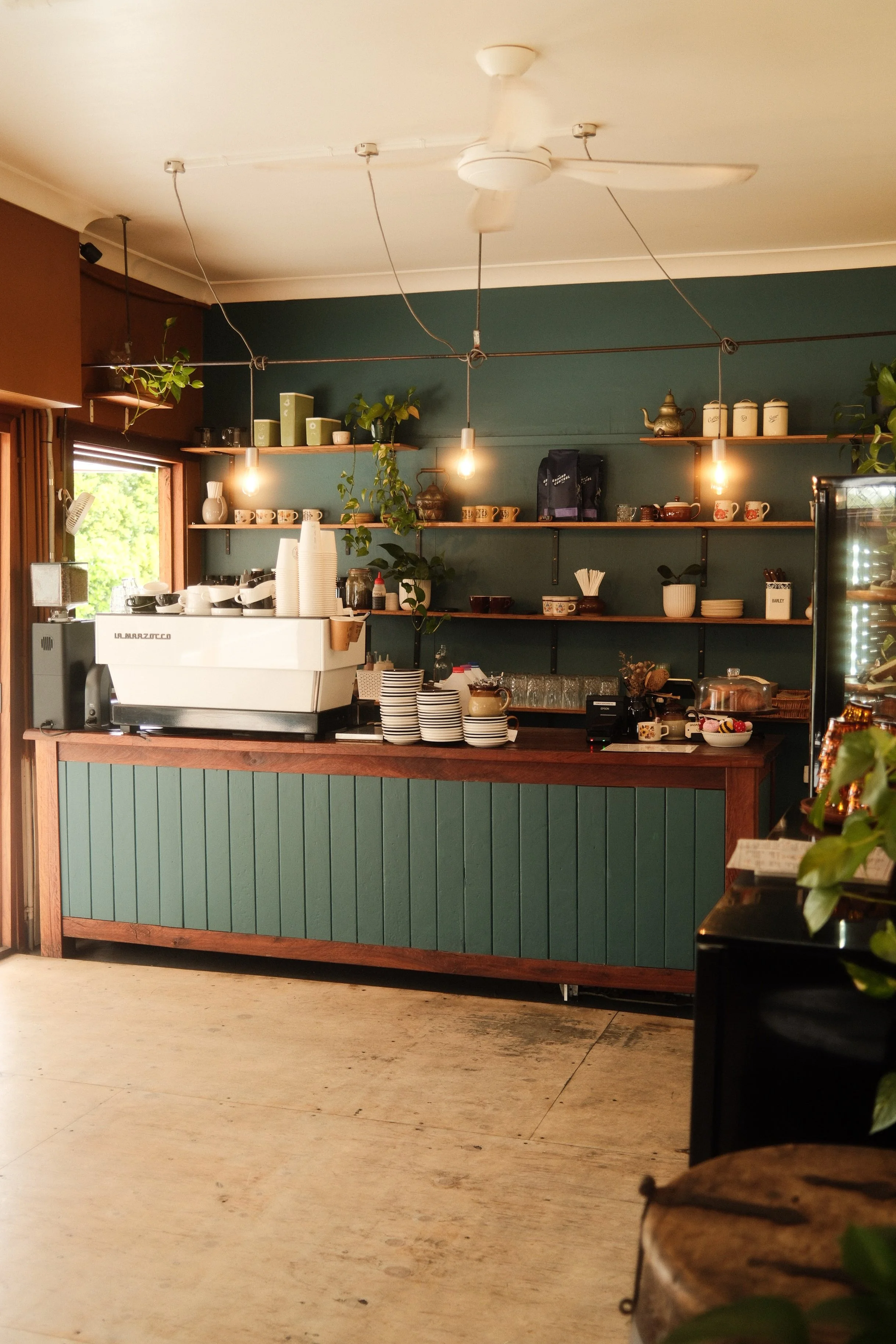 Inneneinrichtung eines Cafés oder Restaurants mit grünem Tresen, offenen Regalen mit Tassen und Geschirr, einem Kaffeeautomaten, einigen Pflanzen und Lichterketten an der Decke.