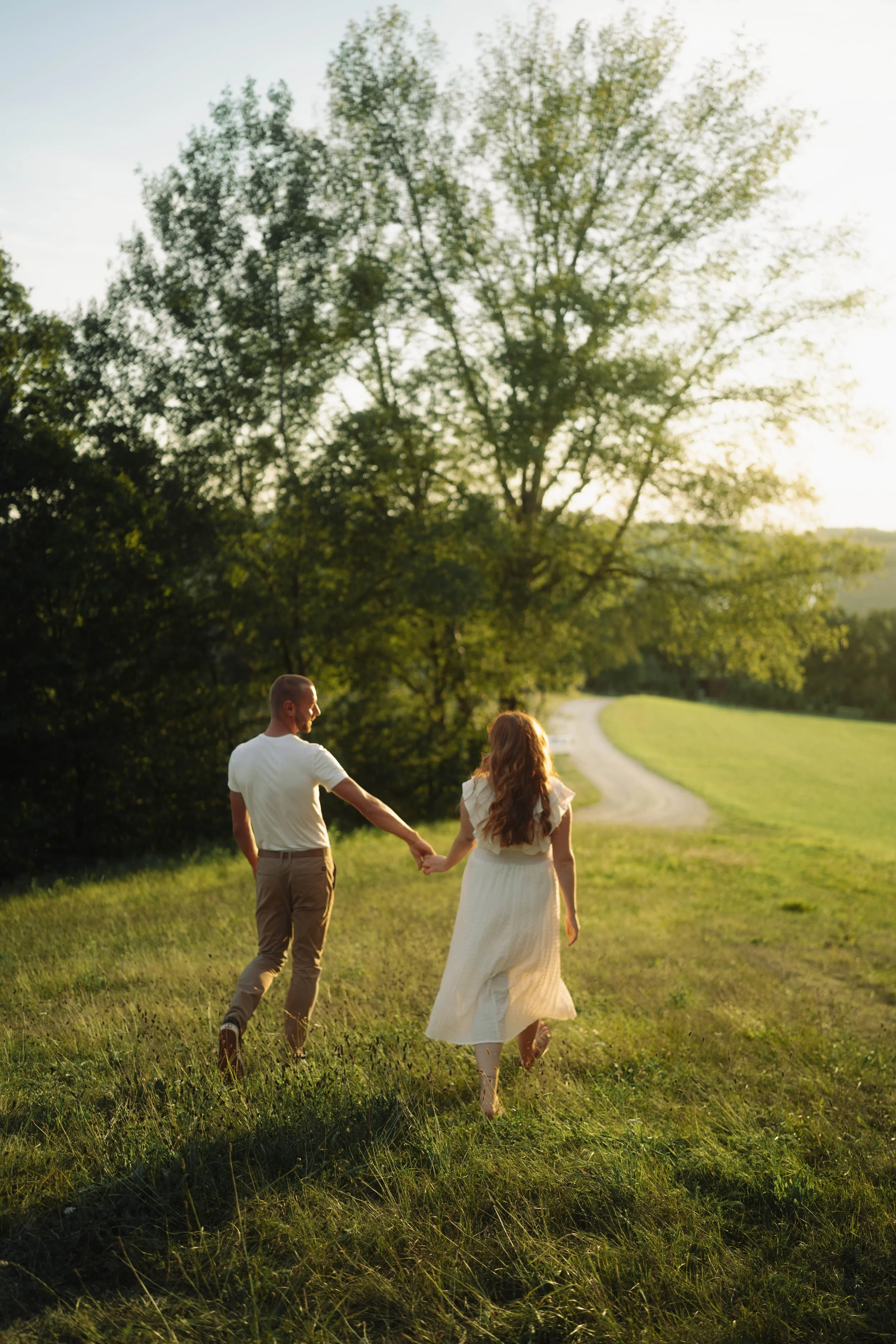 Ein Paar läuft Hand in Hand durch eine grüne Wiese bei Sonnenuntergang, umgeben von Bäumen und einer ruhigen, ländlichen Landschaft.