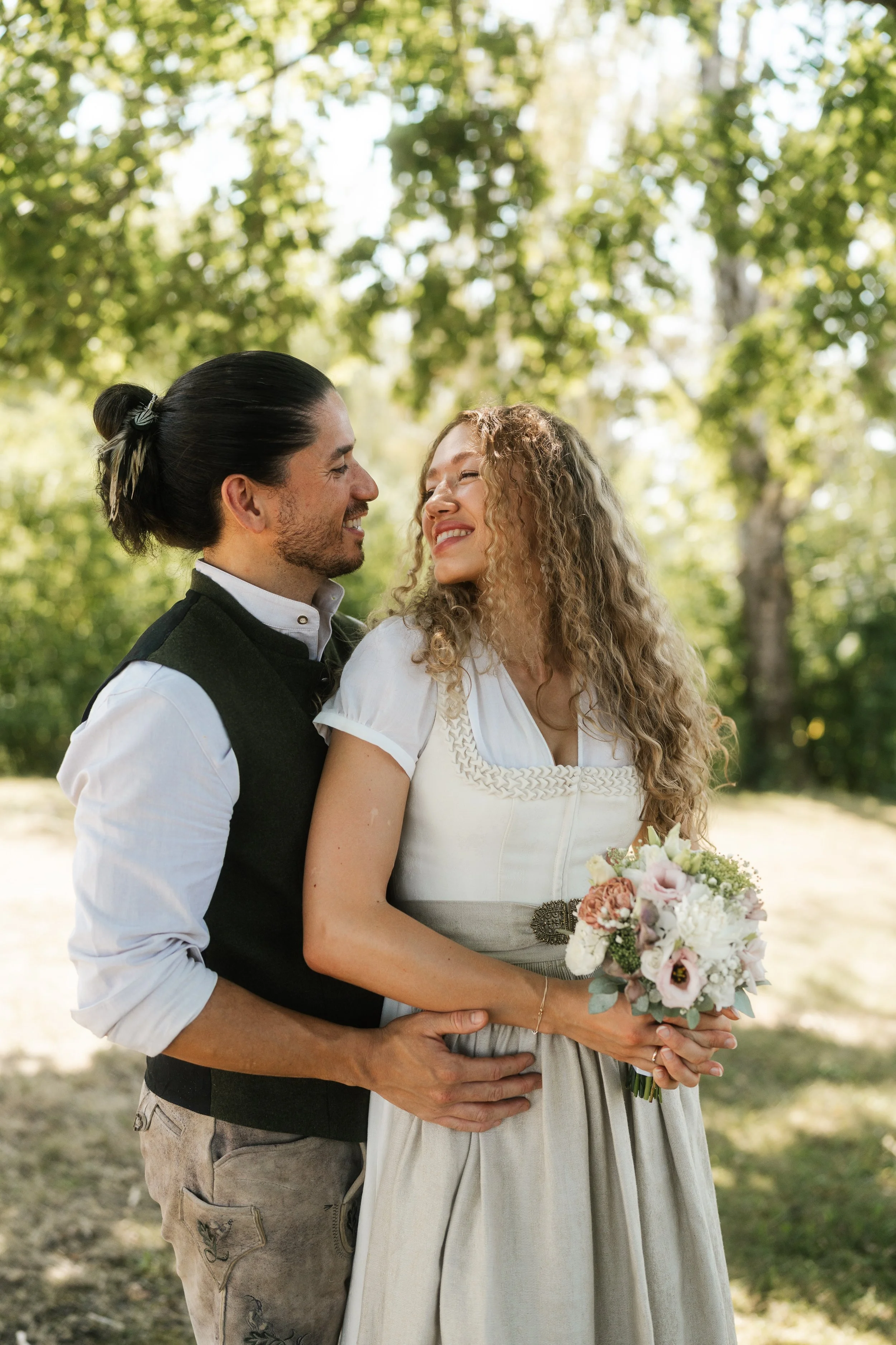 Ein Paar, das bei einer Hochzeit im Freien lächelt. Der Mann hat dunkle, gestylte Haare zusammengebunden, trägt eine weiße Hemd und eine schwarze Weste. Die Frau hat lange, lockige blonde Haare, trägt ein weißes Hochzeitskleid und hält einen Brautstrauß. Der Hintergrund ist mit Bäumen und Sonnenlicht.