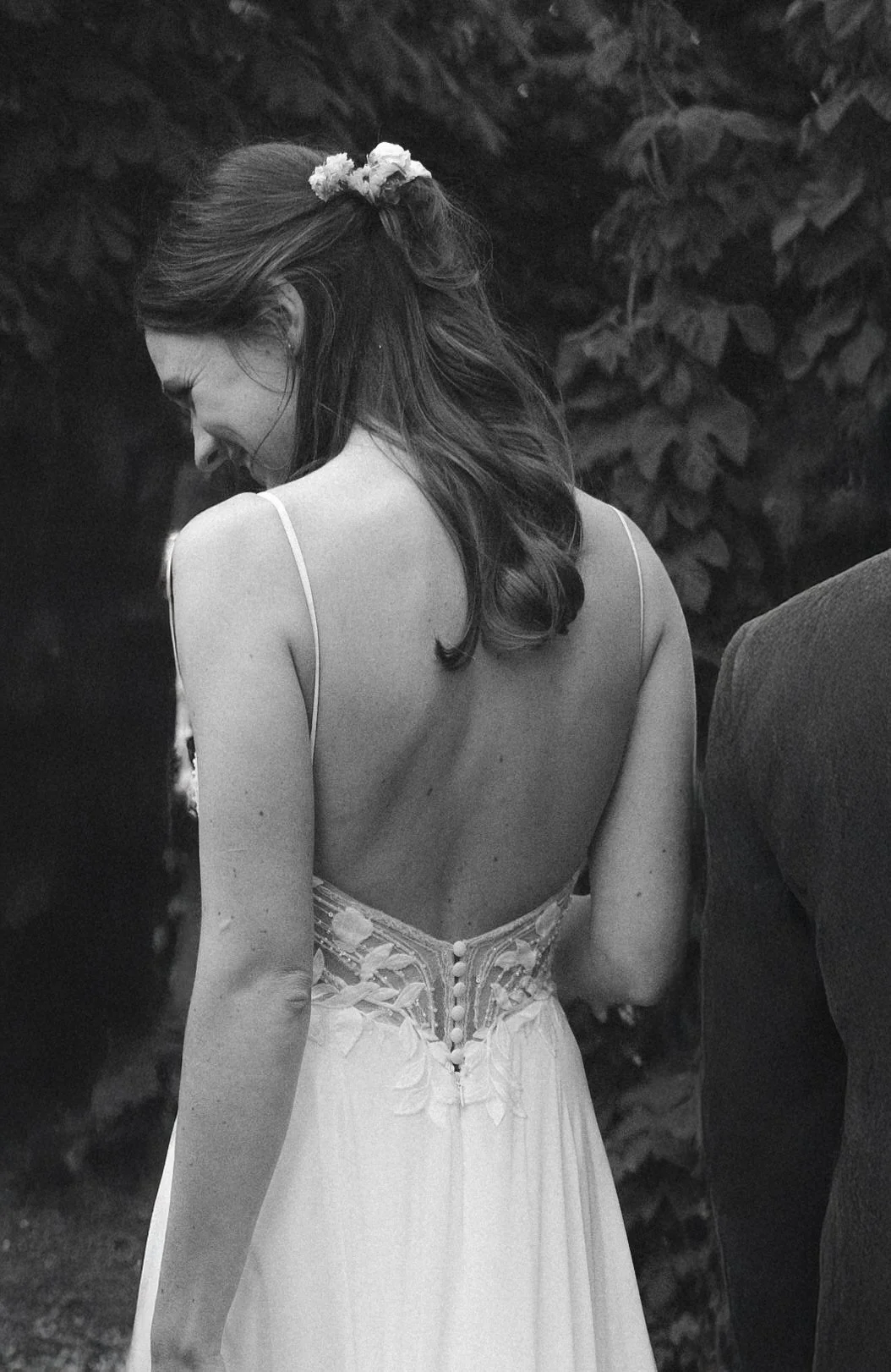 Schwarz-weiß-Fotografie einer lachenden Frau in einem Hochzeitskleid, deren Rücken sichtbar ist, mit einer Blumenkreation im Haar, im Freien vor Büschen.