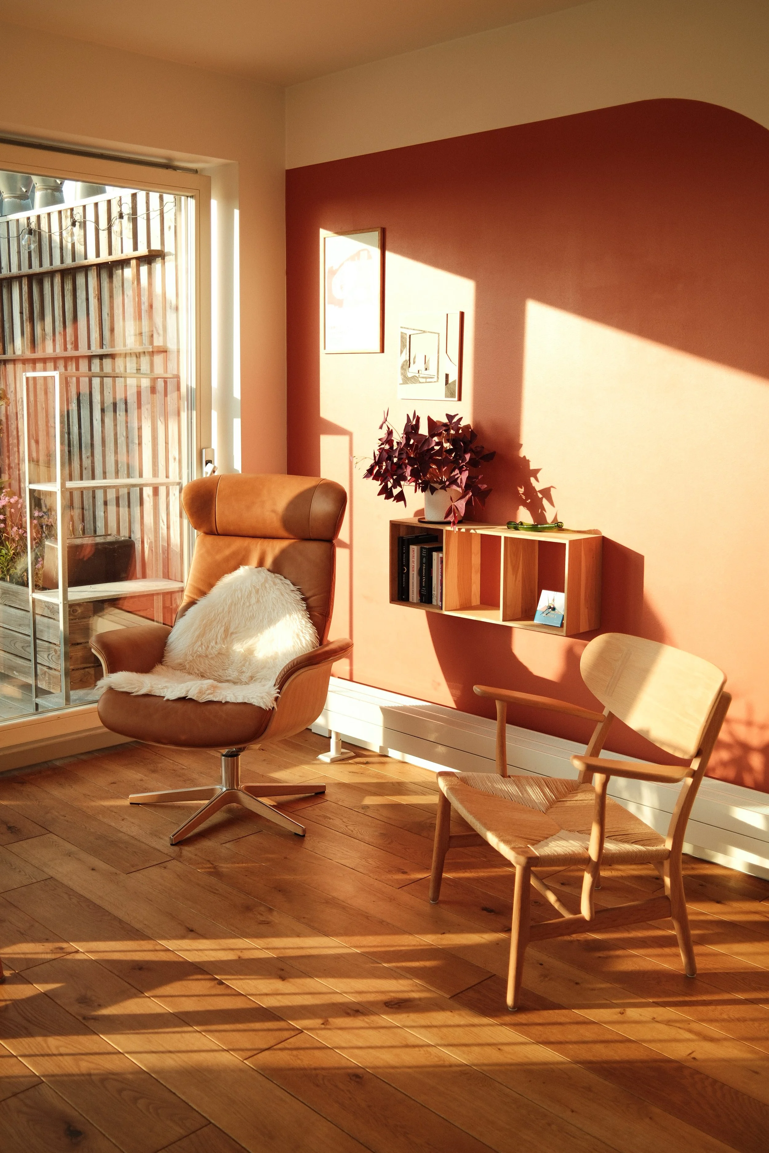 Ein Wohnzimmer mit Holzboden, einem braunen Bürostuhl mit weißen Fell, einer Holzsessel mit Lehne, einem blumigen Topf mit Pflanze, einem Wandregal mit Büchern und Dekorationen, Sonnenlicht wirft Schatten auf die Wand.