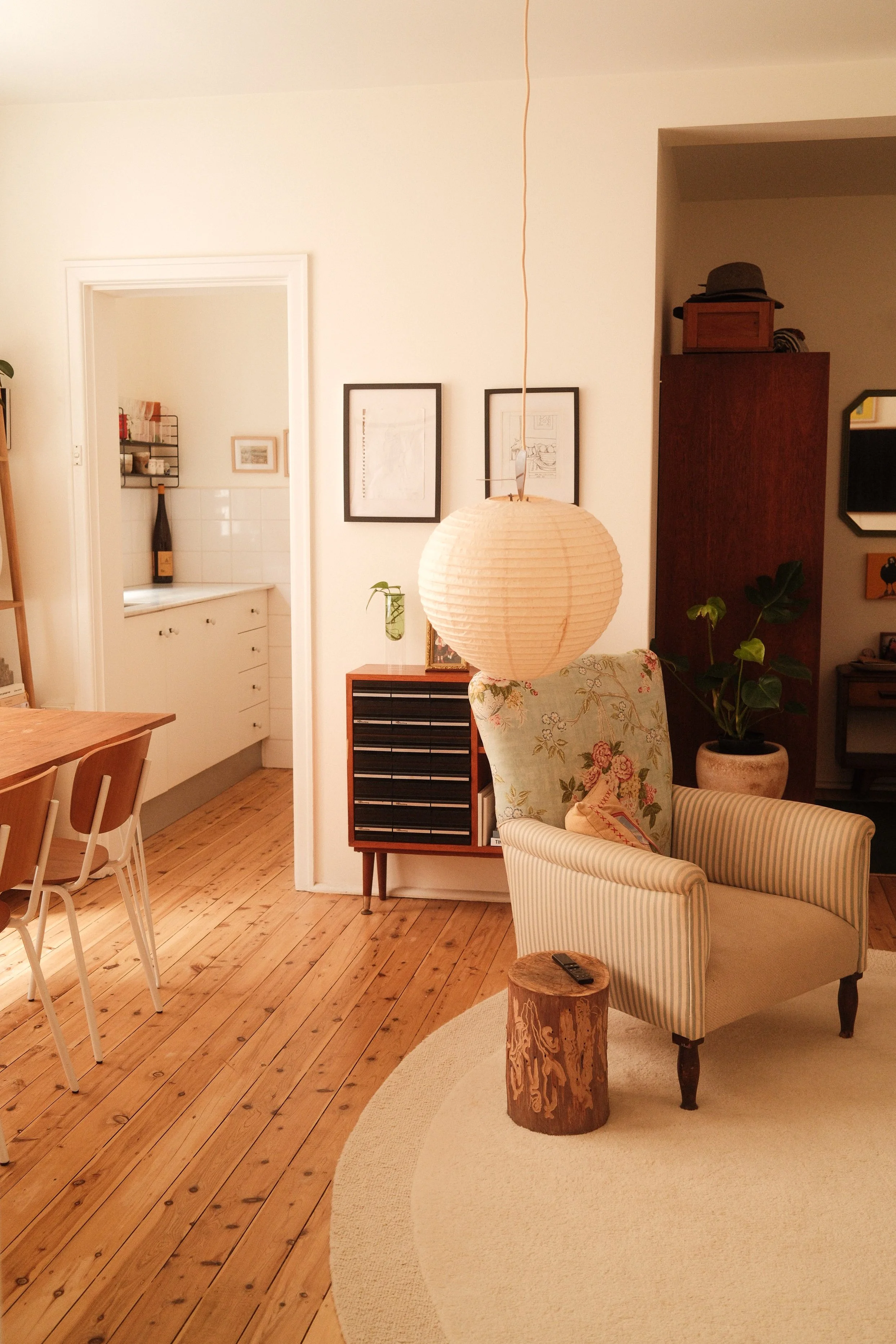 ein Wohnzimmer mit Holzboden, einem gepolsterten Sessel mit Streifenmuster, einem Holzblock-Tisch, einer Stehlampe mit Papierlampenschirm, einem grünen Pflanze in einem runden Topf, mehreren Bildern an der Wand und einer weißen Wand im Hintergrund.