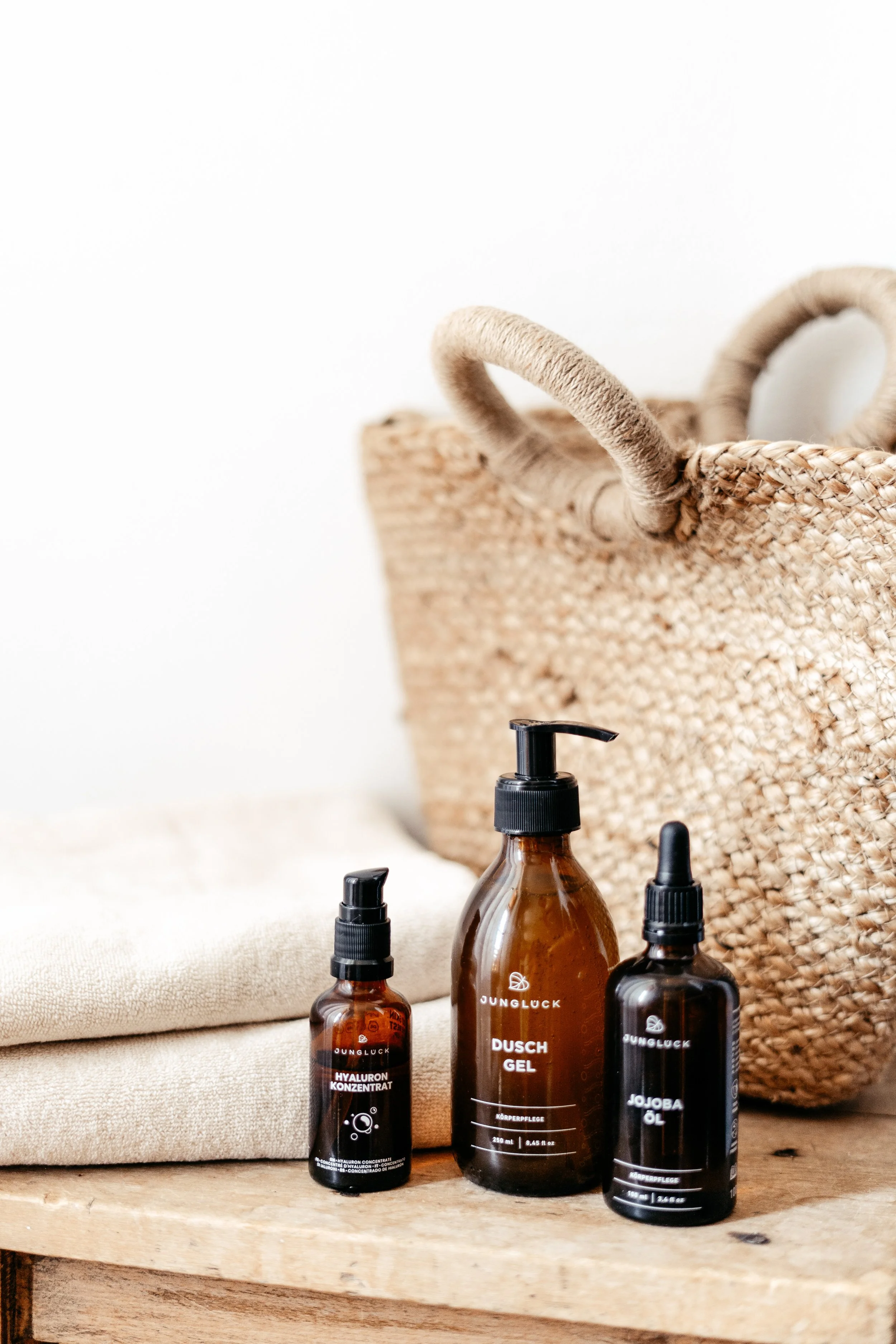 Drei braune Flaschen mit schwarzen Pumpaufsätzen auf einem Holztisch, eine davon mit der Aufschrift 'HYALURON KONZENTRAT', eine mit 'DUSCH GEL' und eine mit 'JOJOBA ÖL'. Im Hintergrund ist eine geflochtene Tasche und ein Handtuch.