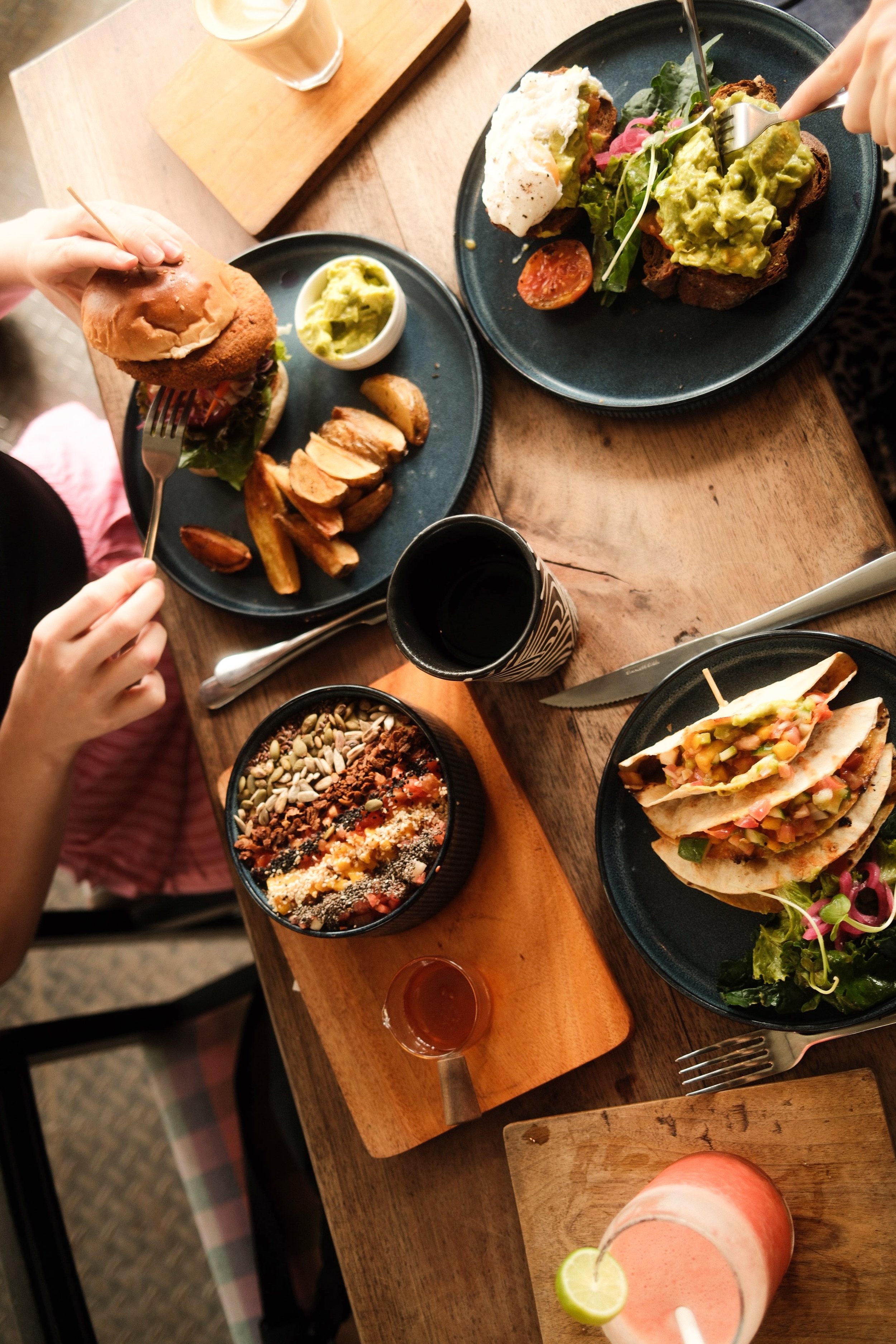 Verschiedene Gerichte, darunter Sandwiches mit Avocado, Salat und Tomaten, eine Bowl mit Müsli oder Porridge, und ein pinker Drink mit Limone auf einem Holztisch in einem Restaurant.