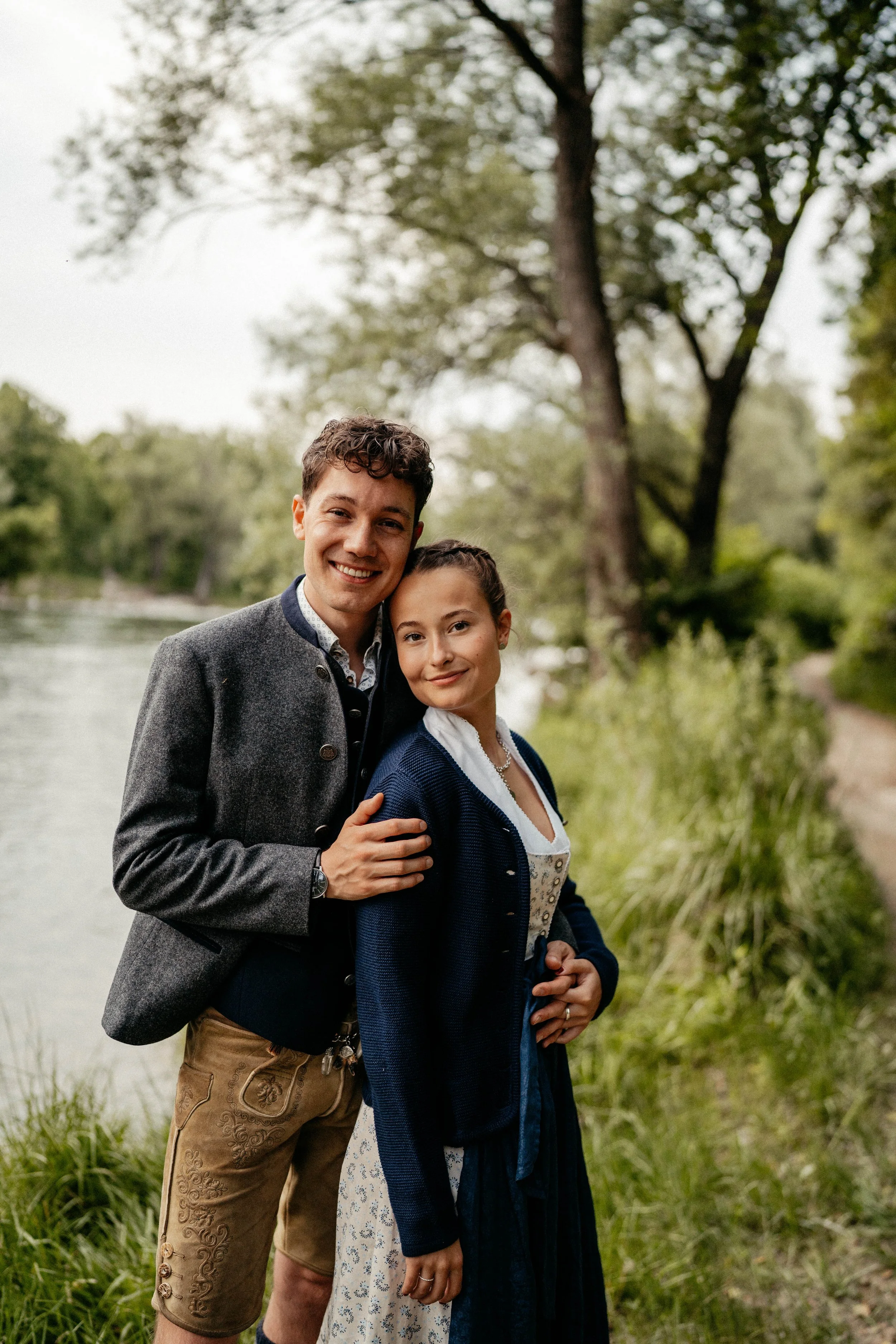 Ein junges Paar, ein Mann und eine Frau, stehen eng beieinander am Flussufer in einer grünen, von Bäumen umgebenen Landschaft. Beide lächeln freundlich in die Kamera, der Mann trägt eine graue Jacke und braune Lederhosen, die Frau trägt ein traditionelles Dirndl. Es ist ein ruhiger und natürlicher Ort, ideal für einen Spaziergang in der Natur.