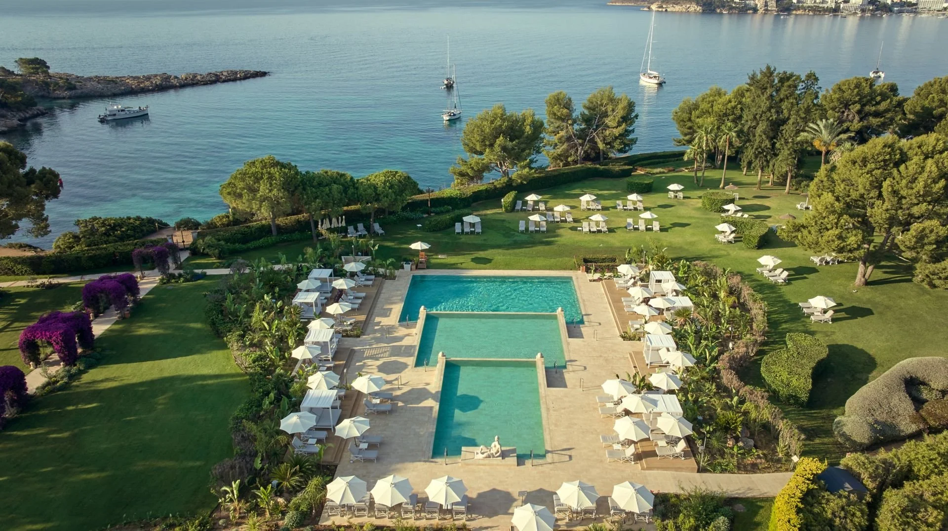 Aerial view of a luxurious resort with a large swimming pool, numerous lounge chairs and umbrellas, surrounded by greenery and trees, overlooking a calm body of water with sailboats and a rocky shoreline in the background.