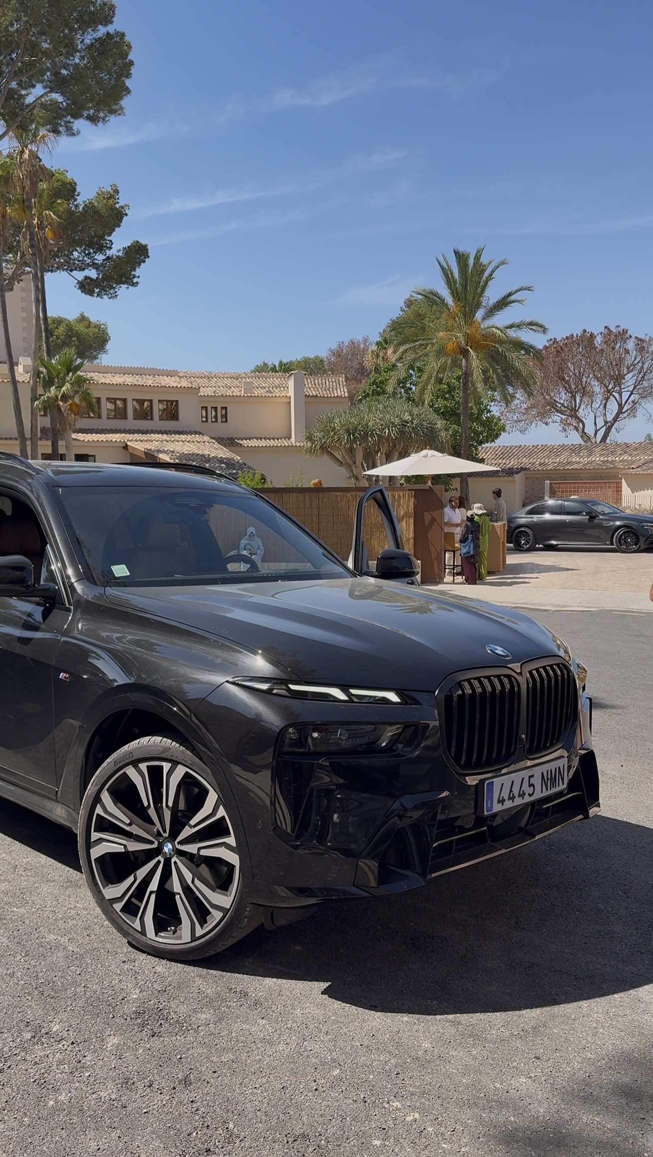 A black BMW SUV parked on a street with a Spanish-style residential neighborhood in the background. The car's front is in view, showing its license plate, and people are seen gathering near a small outdoor stand with an umbrella. The scene features palm trees and other vegetation under a clear blue sky.