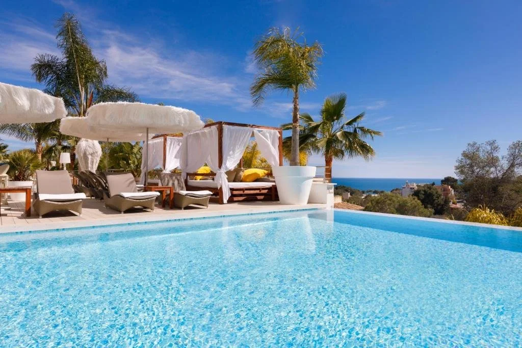 Luxury swimming pool with lounge chairs, umbrellas, and cabanas overlooking a scenic view of trees, coast, and a blue sky.