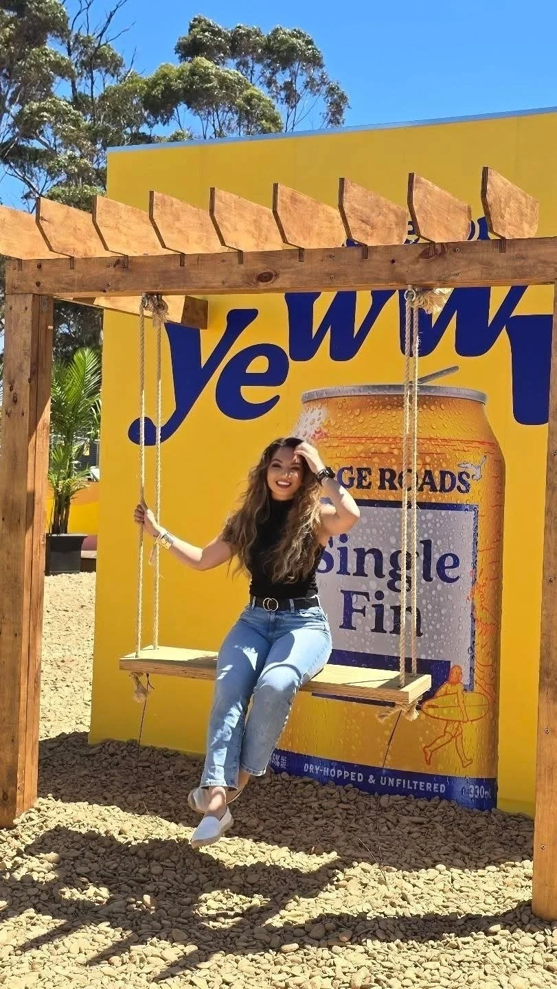 A smiling woman with long hair wearing a black top and jeans sitting on a wooden swing at an outdoor promotional display for a beer brand.