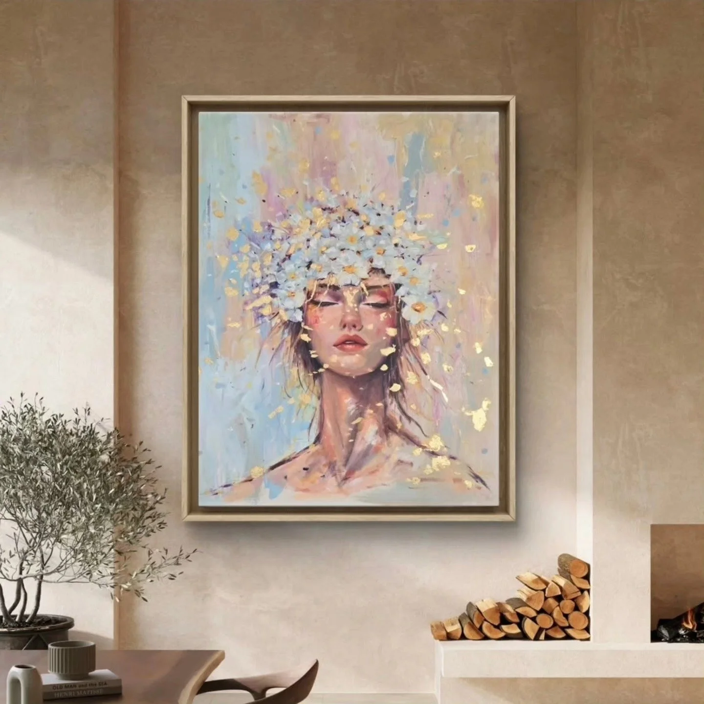 A modern living room with a framed painting of a woman with closed eyes, wearing a crown of white flowers and gold accents, hanging on a beige wall. There is a small potted olive tree and a table with a cup, book, and small decorative items, with a f