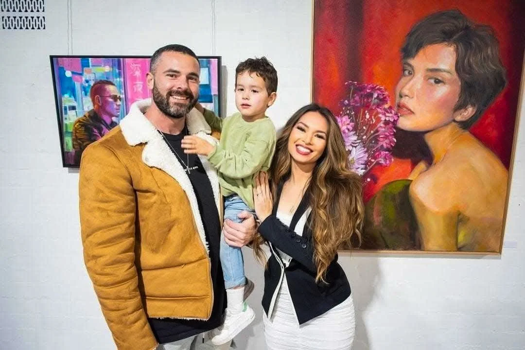 A family of three posing in front of two colorful paintings at an art gallery. The father has a beard and is wearing a tan jacket with a white collar, holding a young boy. The young boy is dressed in a green long-sleeve shirt and jeans. The woman has
