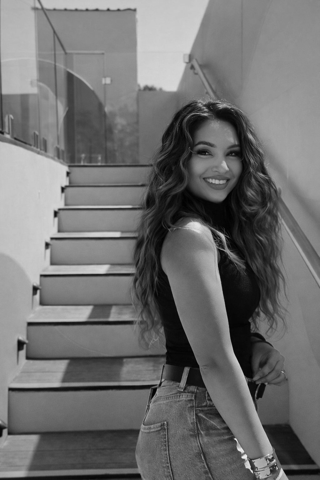 A smiling woman with long wavy hair standing on stairs outdoors, dressed in a sleeveless top and jeans.