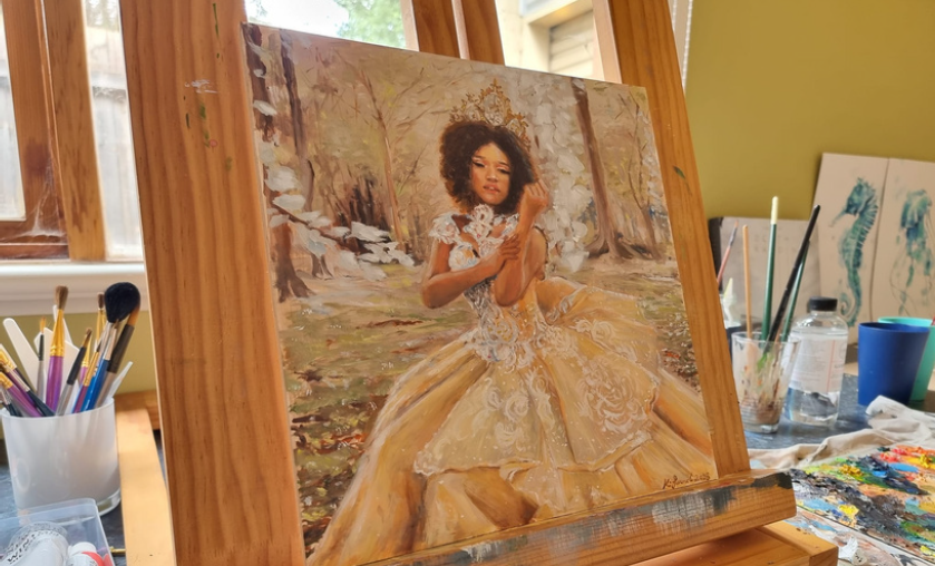 Painting of a woman with curly hair in a yellow dress, sitting outdoors among trees, on an easel in an art studio.