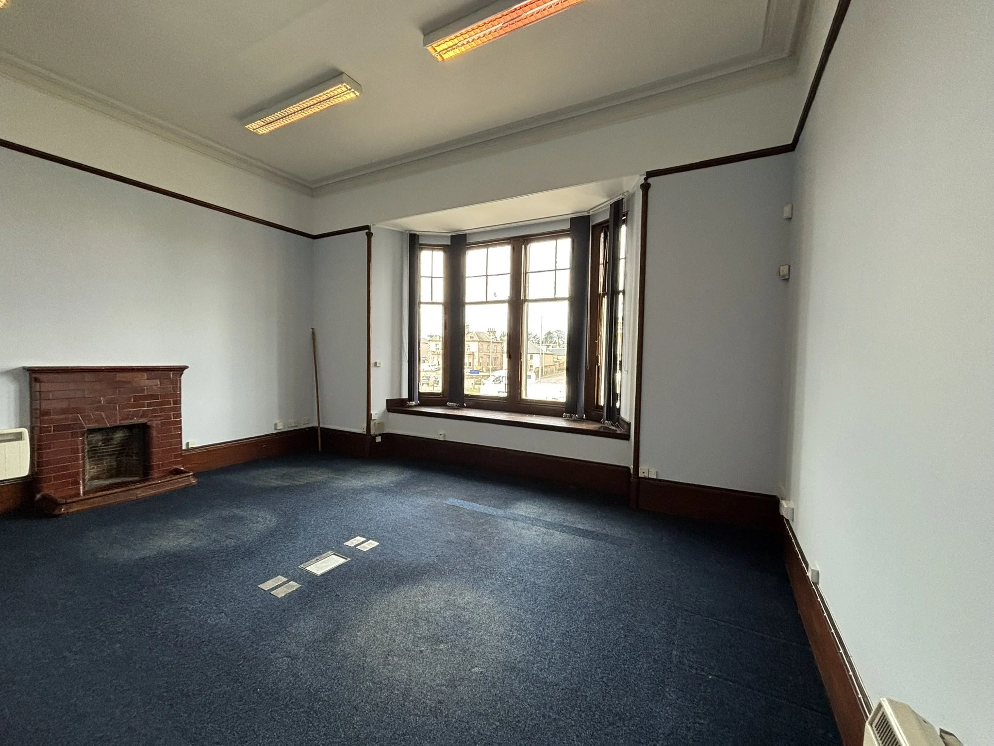 A room with a large window, a brick fireplace, and blue carpeted floor. There is wood trim along the bottom of the walls, and ceiling lights are turned on.