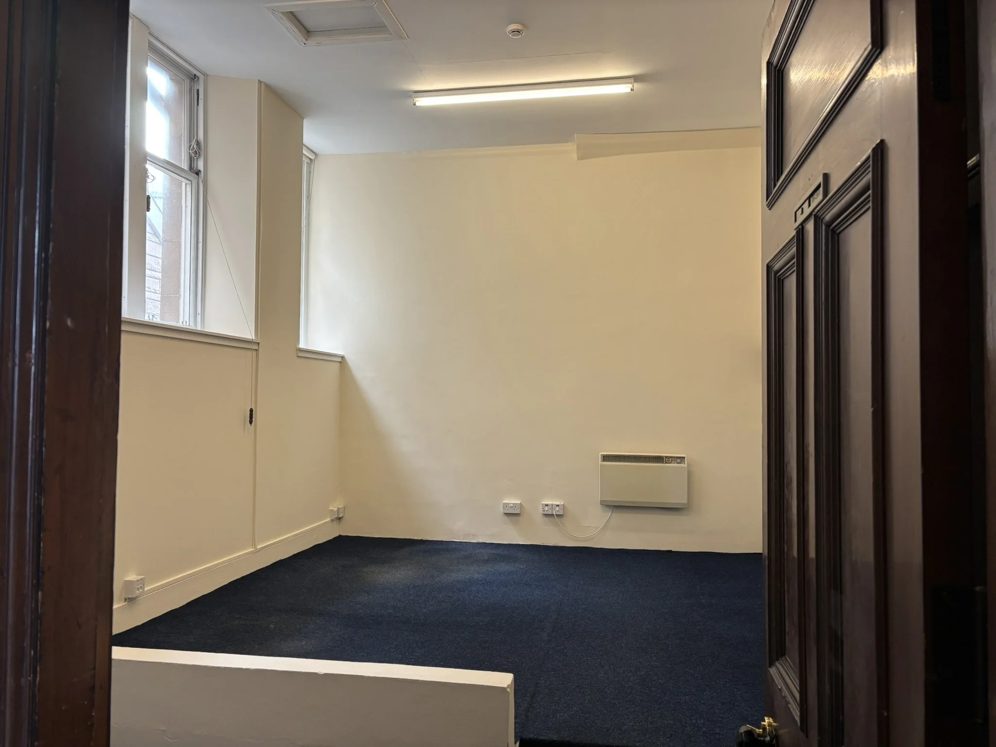 Empty room with cream-colored walls, two windows, a black carpeted floor, and wooden doorframes.