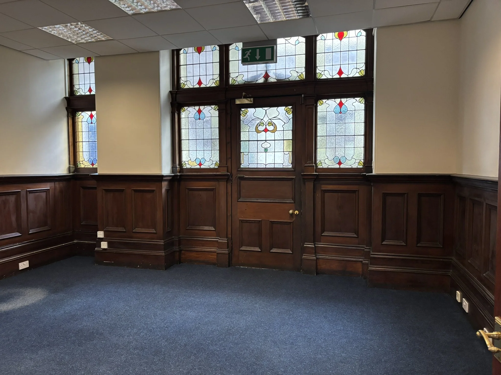 Interior of a room with stained glass windows, dark wooden paneled walls, blue carpet, and multiple electrical outlets.
