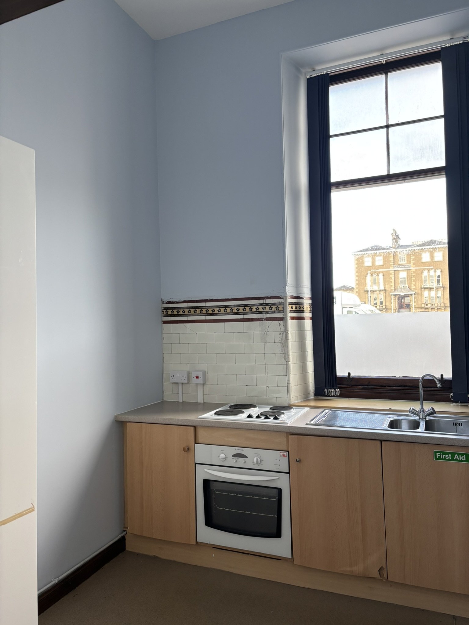 Kitchen with wooden cabinets, a built-in oven, a countertop with a four-burner stove, white tiled wall, a sink, and a window with blue curtains showing a building outside.