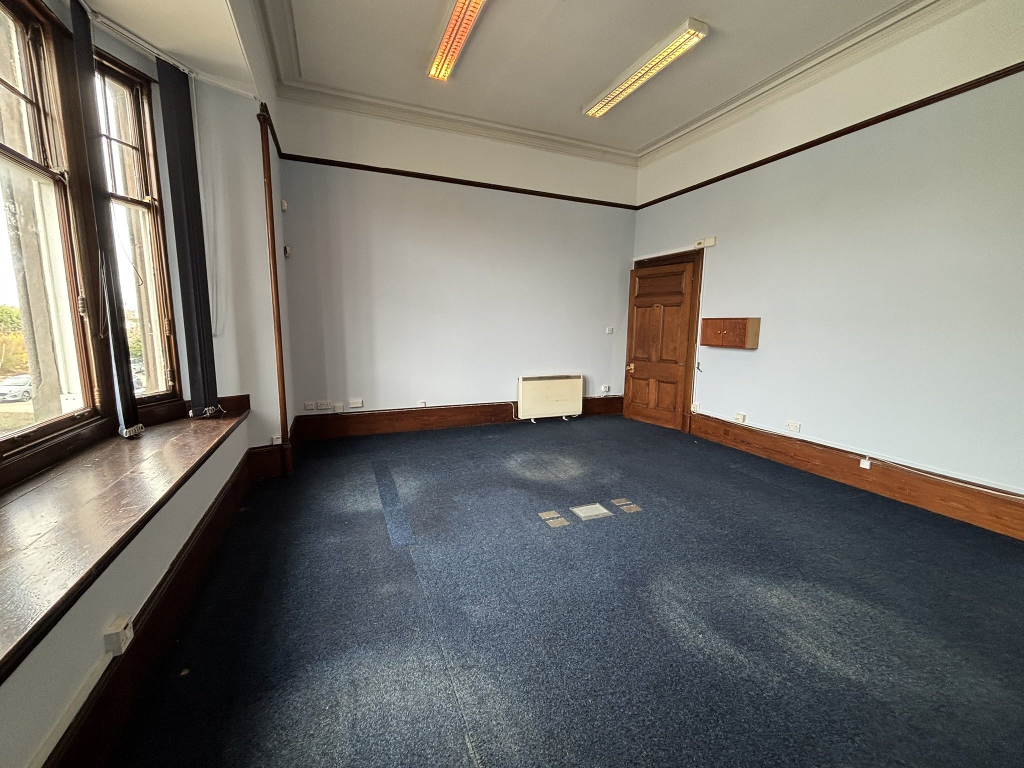 Empty room with large windows on the left, white walls, wooden window and door frames, a baseboard heater, and a blue carpeted floor.