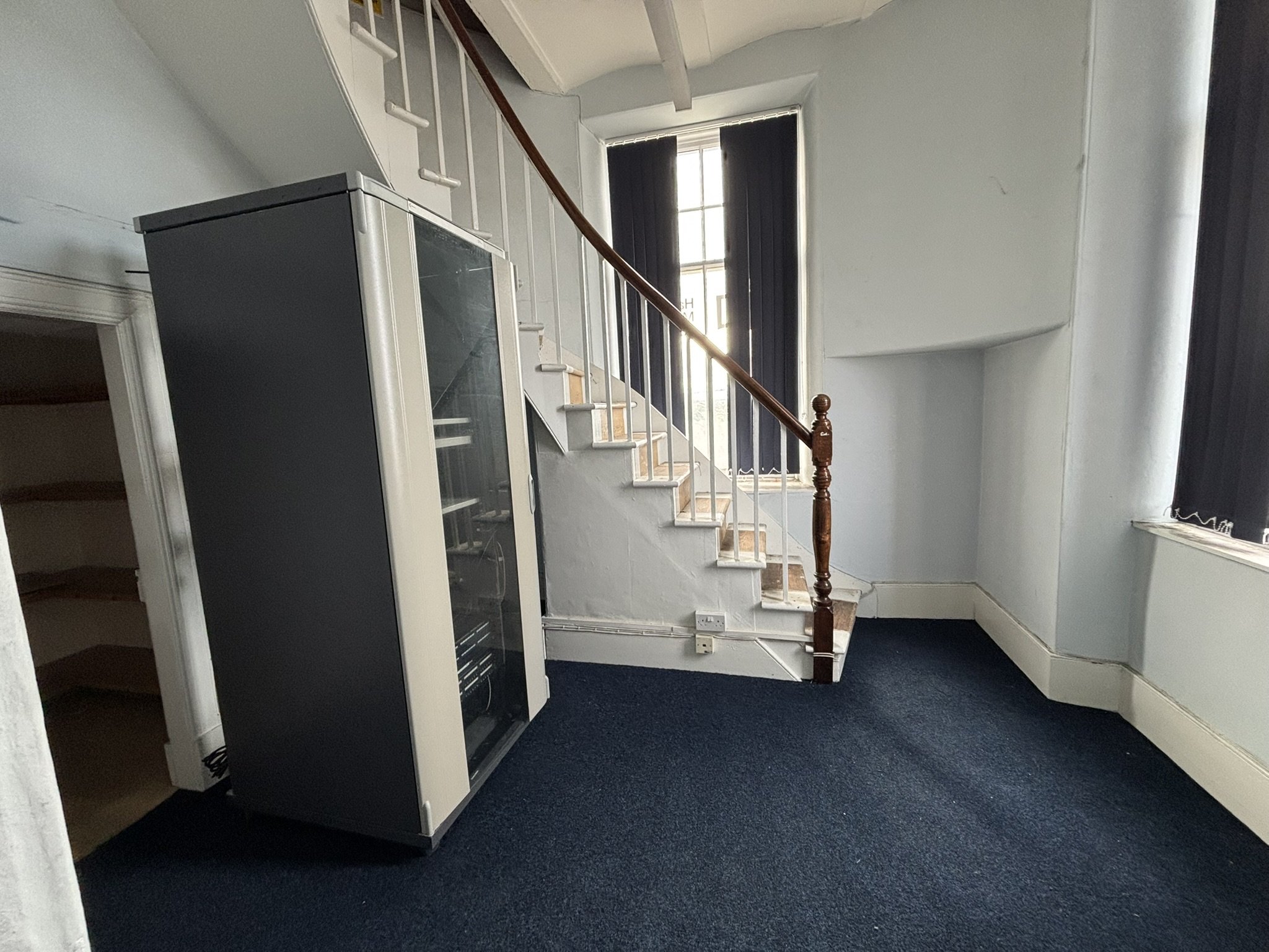 Interior of a house with carpeted floor, staircase with wooden handrail, and a black vending machine or refrigerator near a doorway. There is a large window with black blinds letting in natural light.