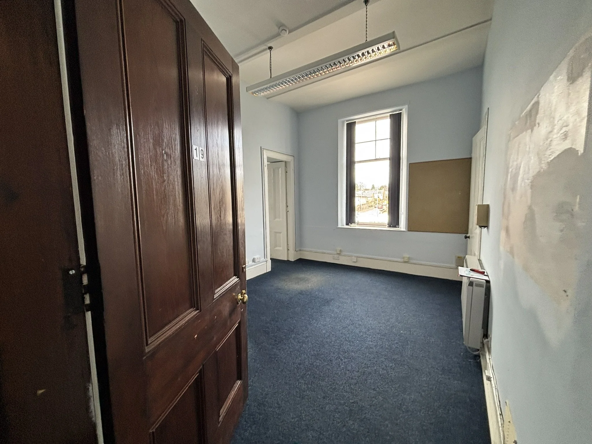 An empty room with white walls, a window, a dark blue carpet, and a large wooden wardrobe.