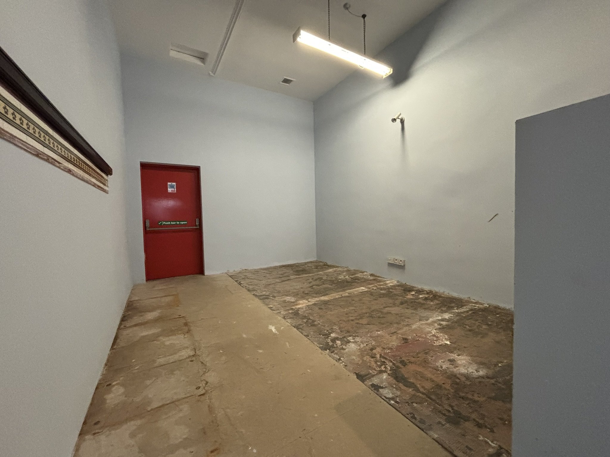 An interior room undergoing renovation with unfinished flooring and white walls. There is a red door with a push bar and a small window, a wall-mounted security camera, a fluorescent ceiling light, and a partially covered window on the left. The floo