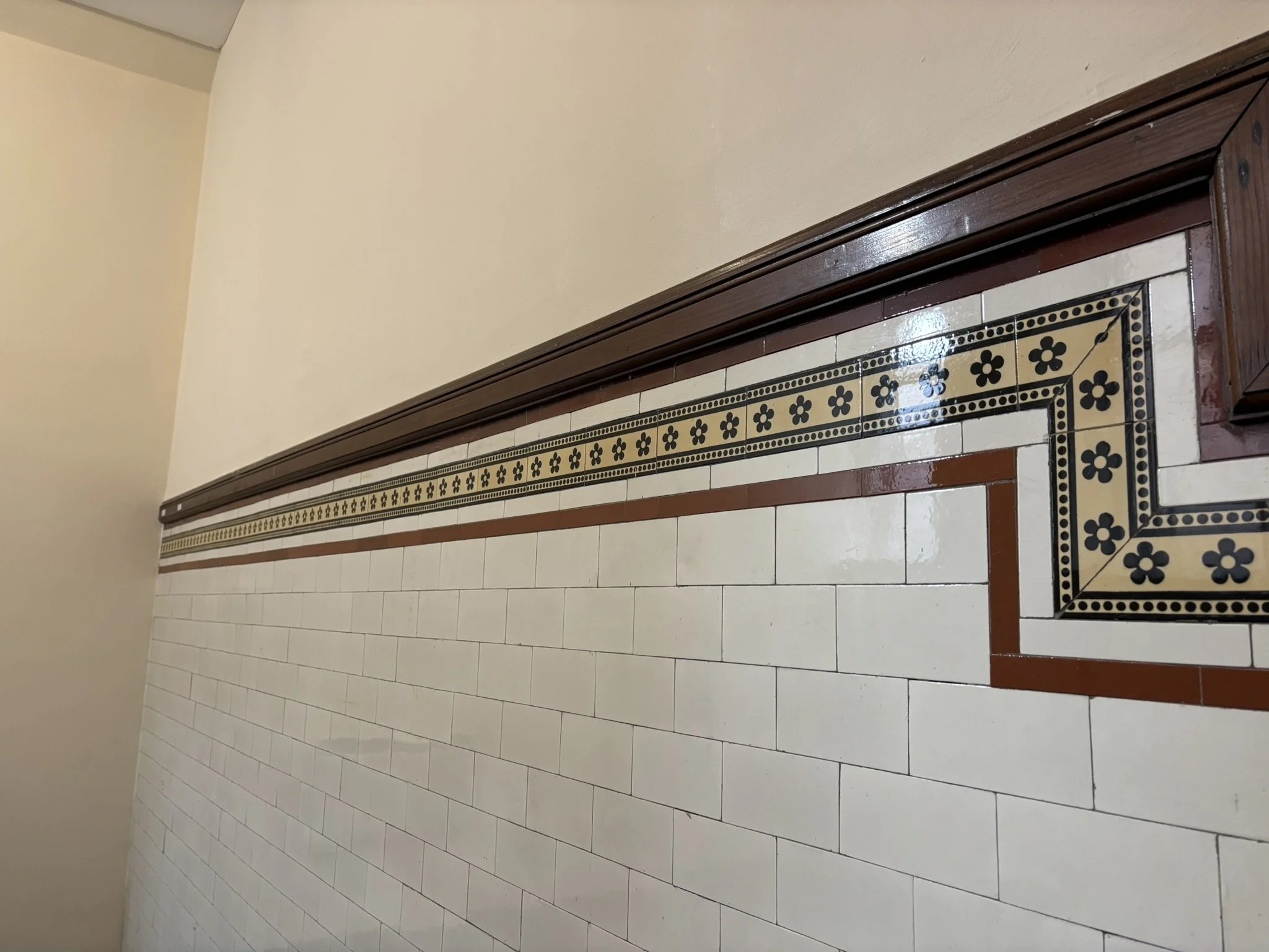 Image of a tiled wall with decorative border tiles, beige and brown paint on the upper part of the wall, and a wooden frame trim.