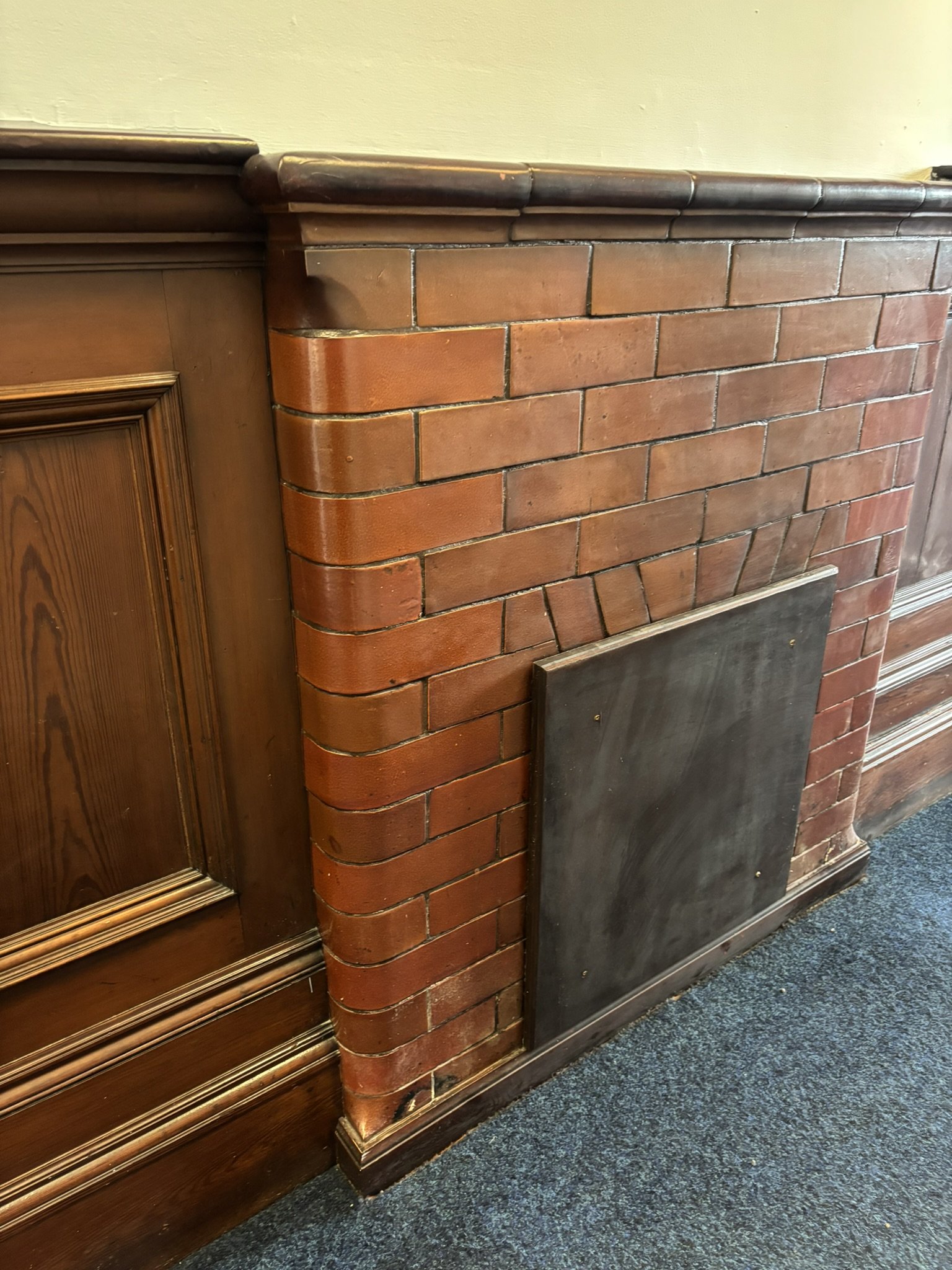 Close-up of a brick fireplace with a wooden mantle and paneling on the adjacent wall, with a dark baseboard and carpeted floor.