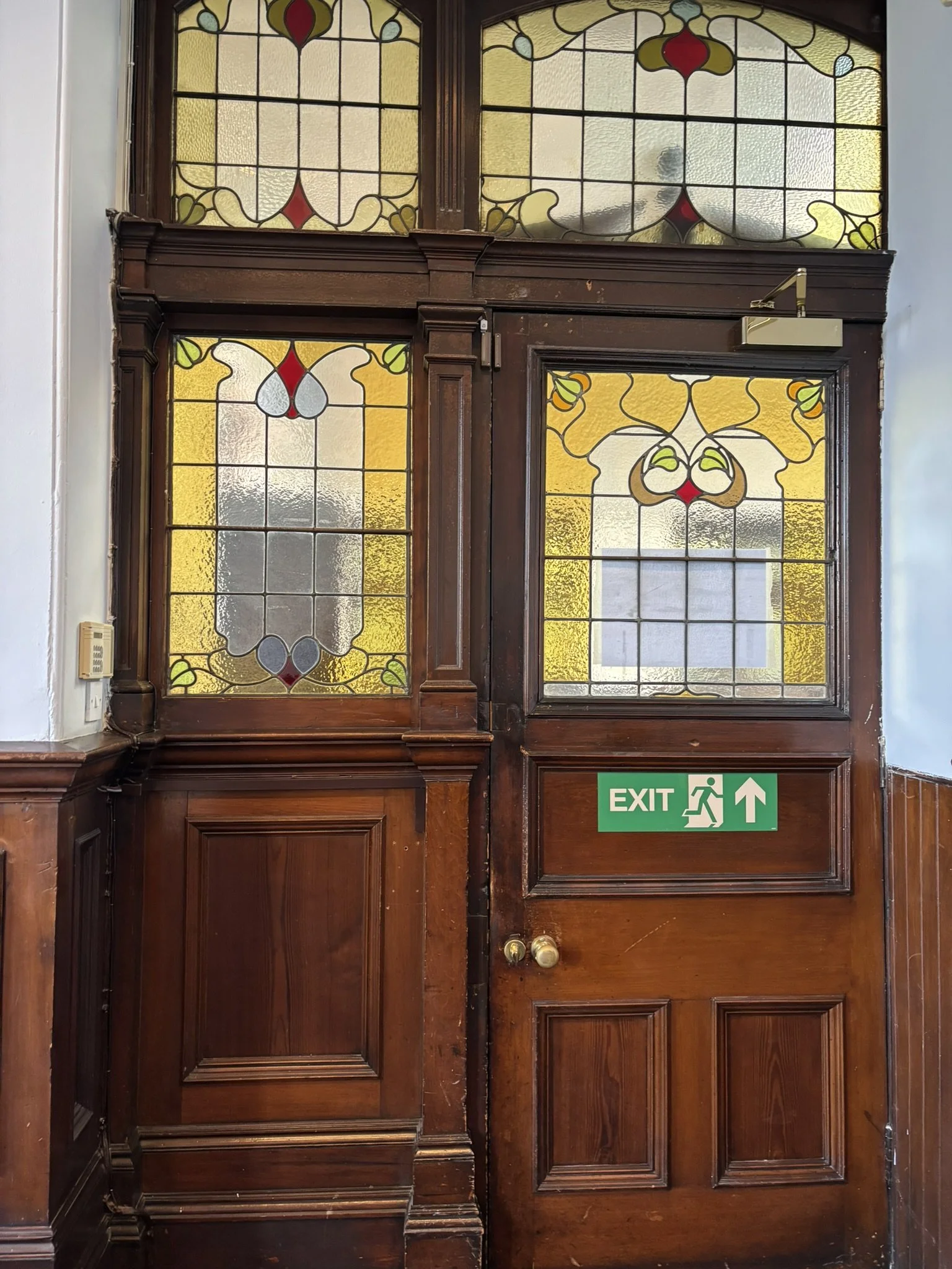 Wooden door with stained glass windows above, featuring geometric and floral patterns in yellow, red, gray, white, and green; green exit sign with arrow on lower panel.