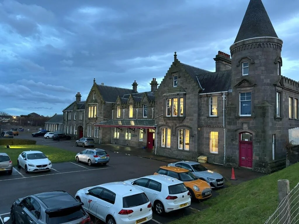 A large castle-like building with a turret, lit windows, and a parking lot with several cars in the foreground during dusk or early evening.