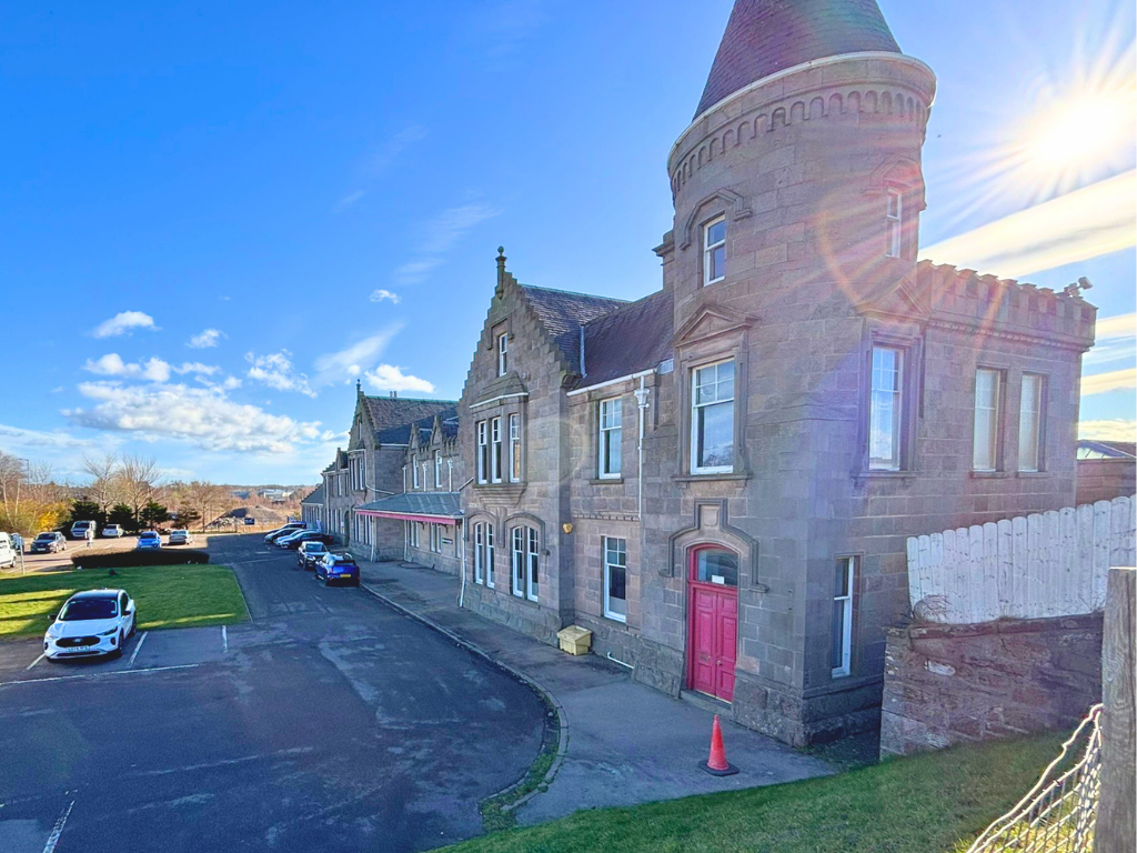 A historic stone building with a conical tower, pink door, and a parking lot in front, under a bright blue sky with some clouds and the sun shining.